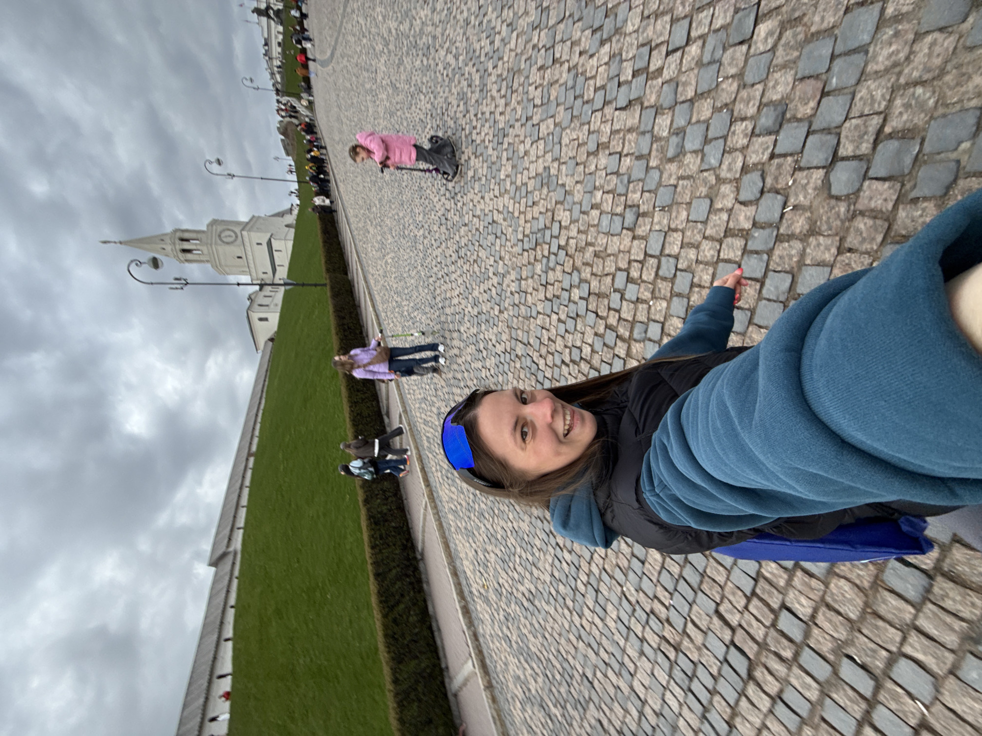 The image is a selfie taken outdoors, likely in Kazan, Russia. In the foreground, a woman with shoulder-length brown hair and wearing blue sunglasses, a black vest, and a blue sweatshirt is looking directly at the camera with a smile. The background features a cobbled stone street leading to a green lawn area bordered by a low hedge and a white stone wall. Behind the lawn, there is a tall white tower with a clock face. Several people are visible in the background, including a child on a scooter and other pedestrians. The sky is overcast. The perspective is somewhat tilted.