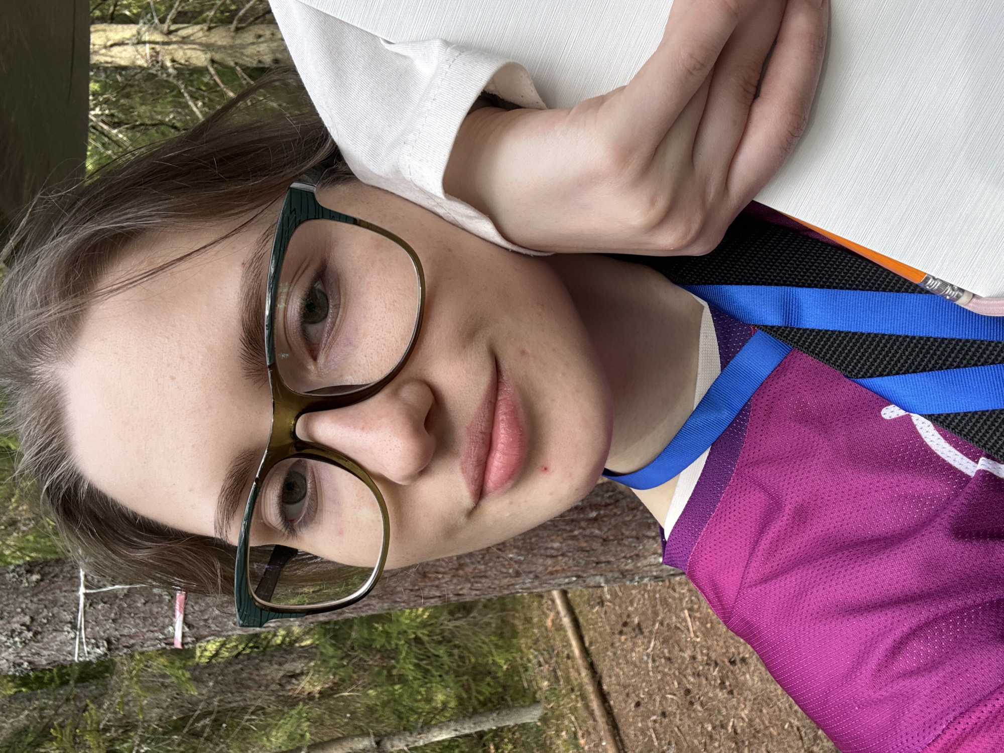 A young woman with glasses is pictured from a close perspective outdoors. She has light skin, light brown hair, and is wearing glasses with dark green rims. She is wearing a white shirt, a pink and black mesh shirt, and a blue lanyard. She is holding a white object with her hand. The background features trees and ground cover, suggesting a forest or park setting. The location is stated as Unknown, Russia.