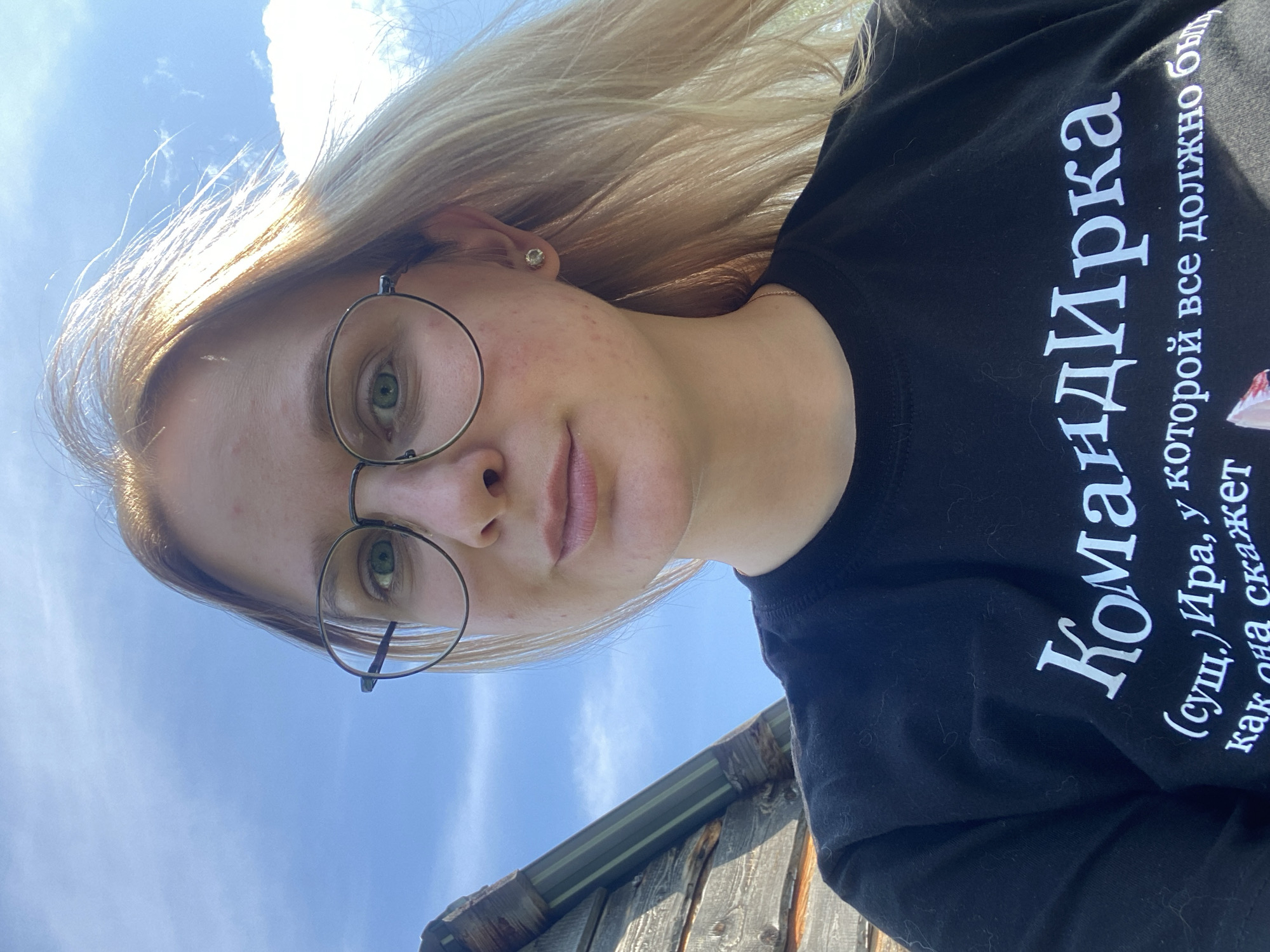 A young woman with blonde hair and green eyes, wearing glasses and a black t-shirt, is captured in a close-up, outdoor selfie. The t-shirt has white Russian text on it. The background consists of a blue sky with some clouds and a portion of a weathered wooden structure, possibly part of a building or shed. The sunlight illuminates her hair from the right. The setting appears to be a rural or semi-rural area, as indicated by the natural lighting and the rustic wooden element. The image's composition focuses on the woman's face and upper torso. The location is stated to be Kauchuk Stantsiya, Russia.