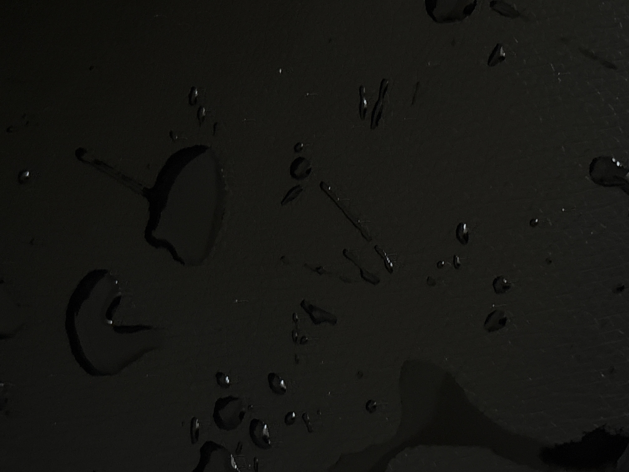The image shows water droplets and spills on a dark, possibly black, surface. The surface appears to have some texture or imperfections. There are no people visible. The drops vary in size from small specks to larger puddles with irregular shapes. No details are available to suggest its location is Kragujevac, Serbia.