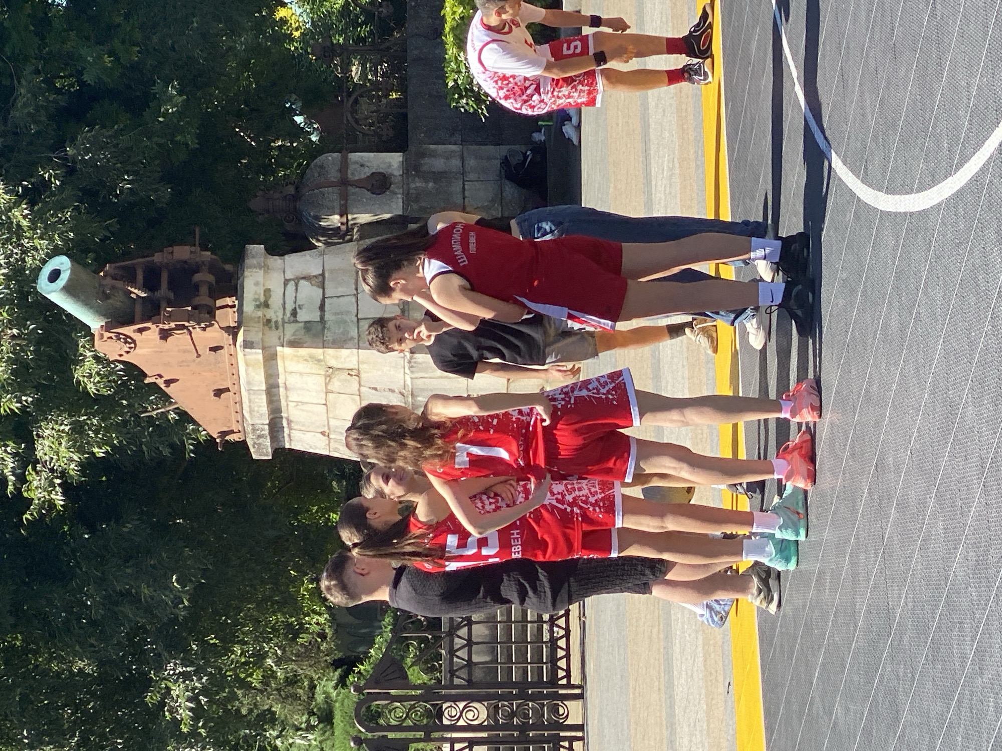 In the city of Pleven, Bulgaria, a group of young people stand on an outdoor court, near a monument featuring a large cannon. The monument is made of stone and weathered metal, with the cannon mounted on top. Behind the monument, trees provide shade. A group of teenagers in red basketball uniforms are gathered in the foreground. One person in a black shirt and shorts stands to the left. Another person in a red and white uniform is bending over on the right. The court is grey with white lines and a yellow border. The overall scene suggests a sports activity or event taking place on a sunny day.