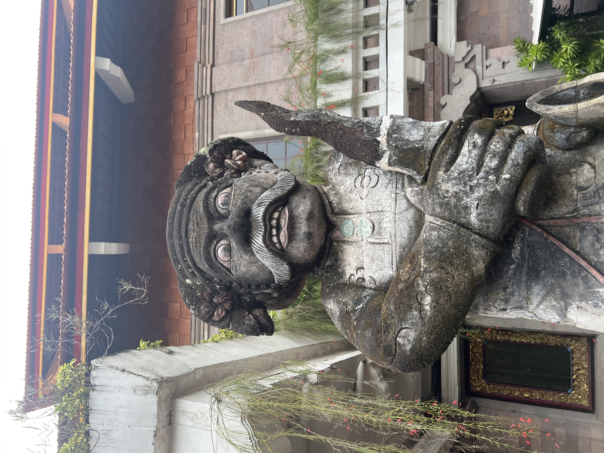 A weathered stone statue of a male figure with an exaggerated expression, featuring bulging eyes, a large, curled mustache, and a wide grin, stands in Celuk, Indonesia. The statue wears a stylized armor or garment with decorative elements. The figure appears to be raising its right arm in a gesture. The backdrop shows a multi-story building with mixed architectural styles, including brick and modern design. Vegetation, including small-leaved plants and red flowers, is visible around the base of the statue. The image is oriented vertically, with the statue dominating the foreground.