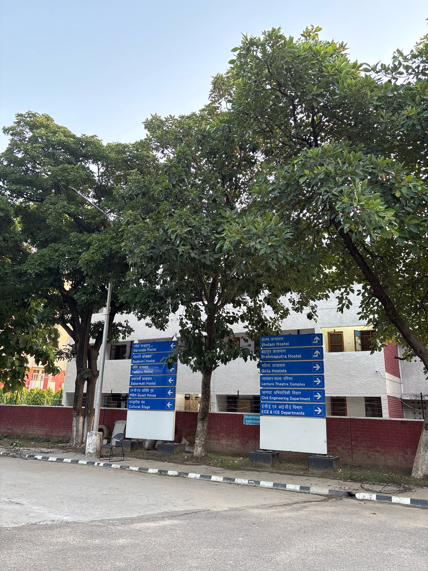 The image shows a street-level view of a building in an outdoor setting. A tall, white building with red brick trim is visible in the background, partially obscured by large, leafy green trees. In front of the building, a multi-directional blue signpost with white text indicates various locations such as "Narmada Hostel," "Godavari Hostel," "Yamuna Hostel," "Sabarmati Hostel," "Jhelam Hostel," "Brahmaputra Hostel," "Girls Hostels," "Lecture Theatre Complex," "Civil Engineering Department," and "ECE & ICE Departments." The signpost is illuminated by a streetlamp. The ground in the foreground is paved, with a curb and a sidewalk. A black metal chair is placed near the signpost. The sky is clear and pale blue.
