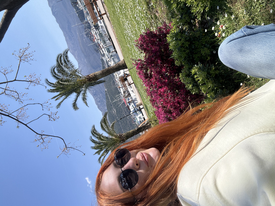 An outdoor, daytime image depicts an individual seated in a park-like setting overlooking a marina in Budva, Montenegro. The subject is a person with long, bright reddish-orange hair, wearing dark sunglasses, a light-colored hooded top, and light blue denim jeans. The person's face is partially visible, oriented towards the left foreground, with a slight smile.
The immediate foreground features green grass dotted with small white flowers. Behind the person, a dense bush with vibrant dark pink/magenta flowers is visible, alongside other green foliage. Overhead, bare tree branches with small buds or nascent leaves extend into the clear blue sky.
The midground reveals a marina with numerous white boats and yachts docked in the water. A paved walkway runs alongside the marina, where several light-colored wooden benches are positioned. At least two tall palm trees stand between the park area and the marina. The background is dominated by large, rocky mountains under a clear, bright blue sky with a single faint contrail visible. The scene is illuminated by strong sunlight, casting visible shadows.