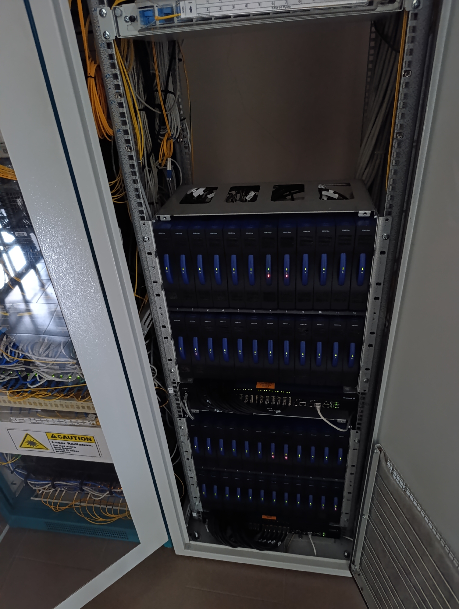 A rack of servers is shown in a room. The servers have blue lights on their front panels. There are cables running throughout the rack. A caution sign for laser radiation is visible on one of the servers. The image appears to be taken in the city of Dubăsari town council, Moldova.