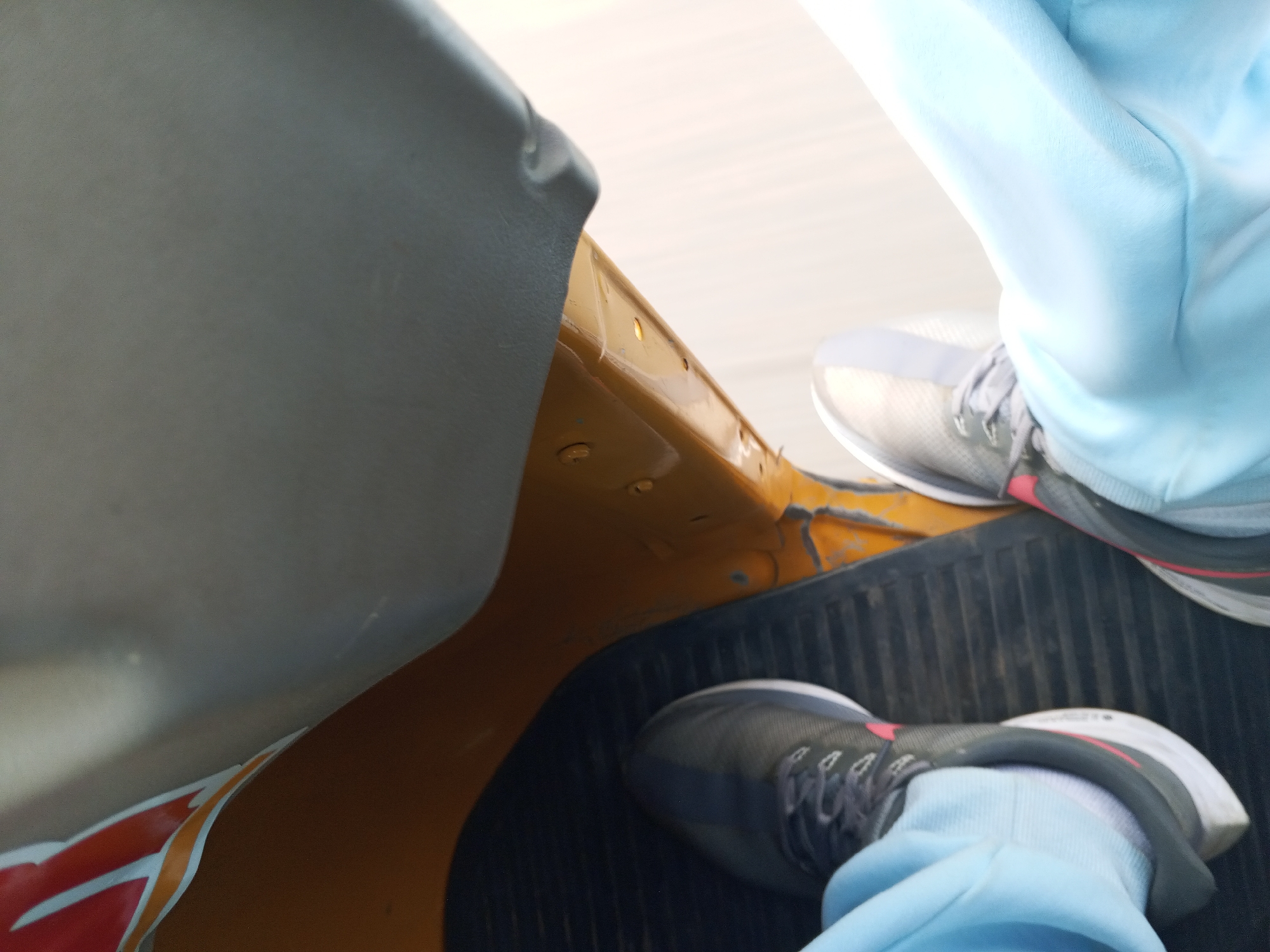 This is a close-up, interior shot of an auto-rickshaw in Karu, Nigeria. Two pairs of feet, clad in light blue sweatpants and athletic shoes, are visible. One pair of feet is positioned on the floor mat of the auto-rickshaw, while the other pair is resting slightly higher, possibly on a seat. The interior of the auto-rickshaw appears to be yellow and grey. The floor mat is black and ribbed. The lighting suggests it is daytime.