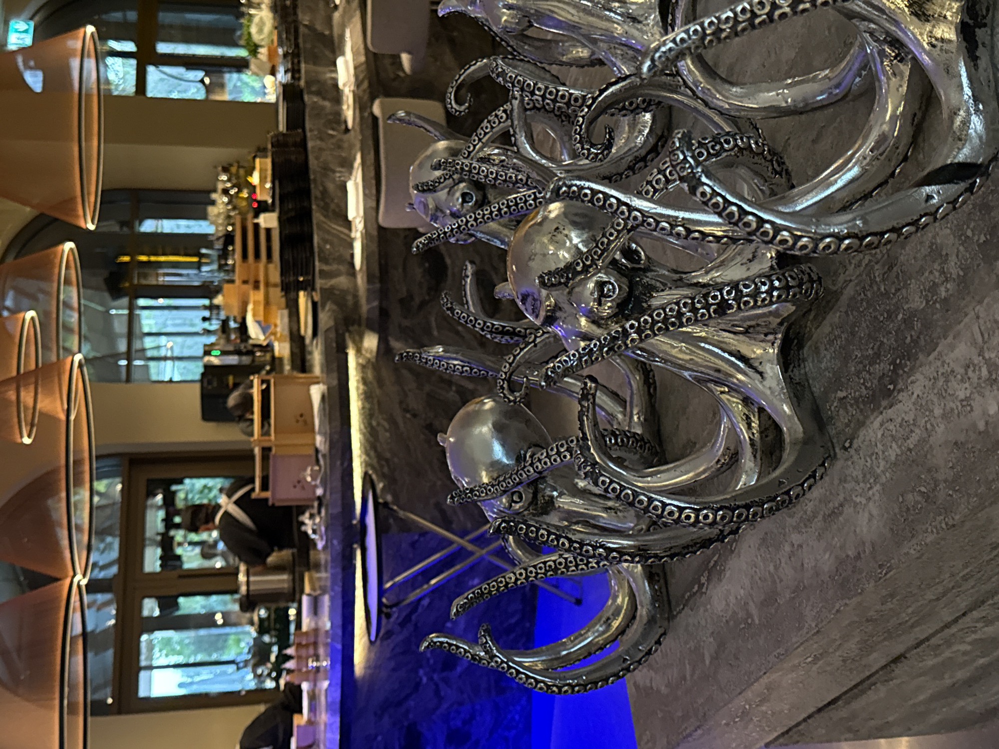 The image shows a close-up of several silver octopus sculptures, with their tentacles intertwined, placed on a concrete surface. In the background, a dimly lit restaurant or bar area is visible. There are tables with white napkins and chairs, a marble counter with various items including stacks of plates and serving ware, and large windows that look out onto a green outdoor space. A chef in a black uniform and apron is partially visible behind the counter. The lighting is a mix of ambient restaurant lighting and a strong blue light emanating from a surface in the foreground. The scene appears to be an interior of a high-end dining establishment. The location is identified as a restaurant in Dubai, United Arab Emirates.