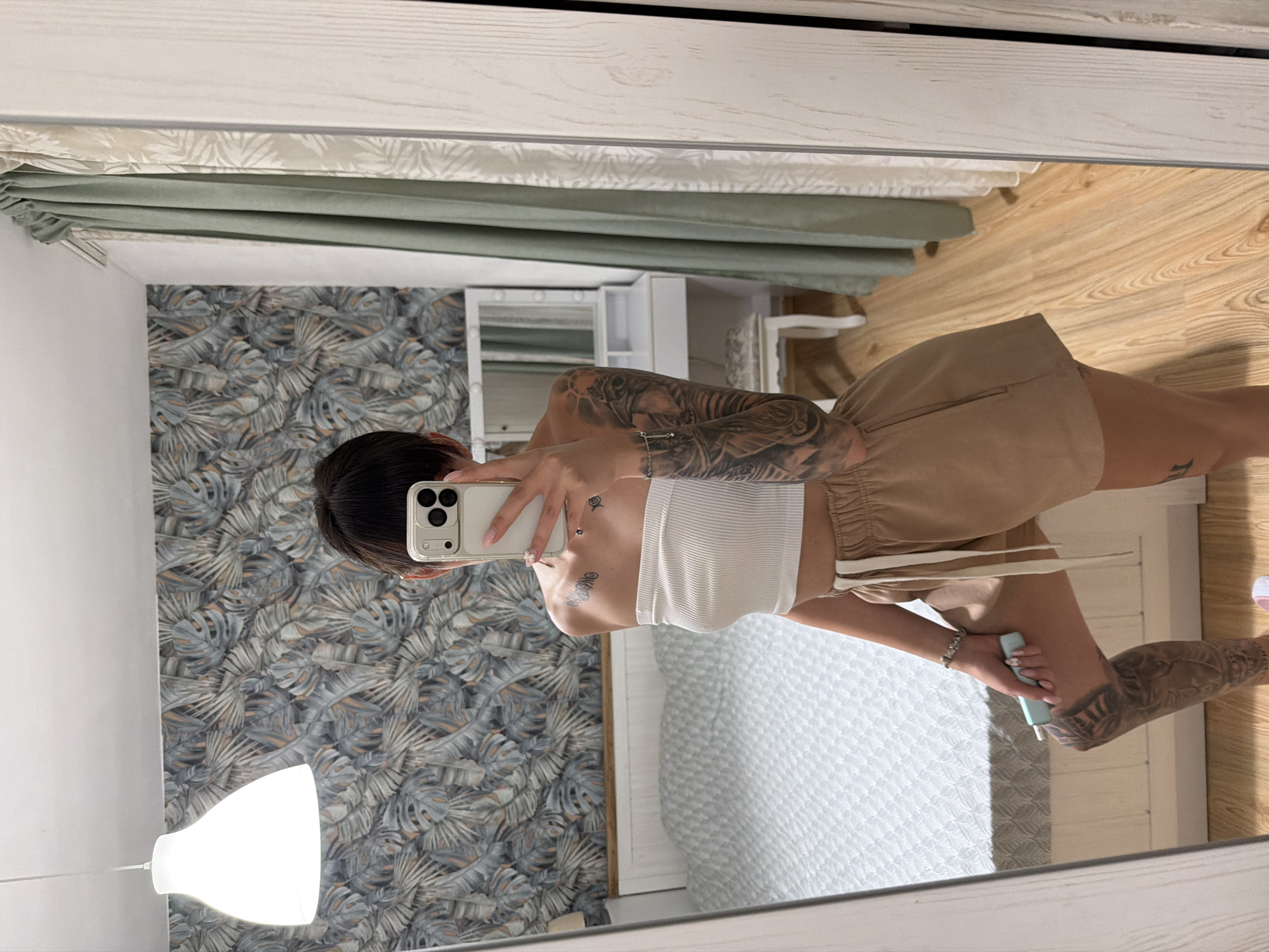 A young woman with short, dark hair is taking a mirror selfie in a bedroom. She is wearing a white strapless top and beige shorts. She has tattoos on her arms and legs. She is holding a light blue object in her right hand. The room has a bed with white bedding, a lamp, patterned wallpaper, and curtains. There is also a vanity with a mirror in the background. The floor is made of light-colored wood. The image does not provide any specific location information, so it cannot be definitively placed in Plovdiv, Bulgaria.