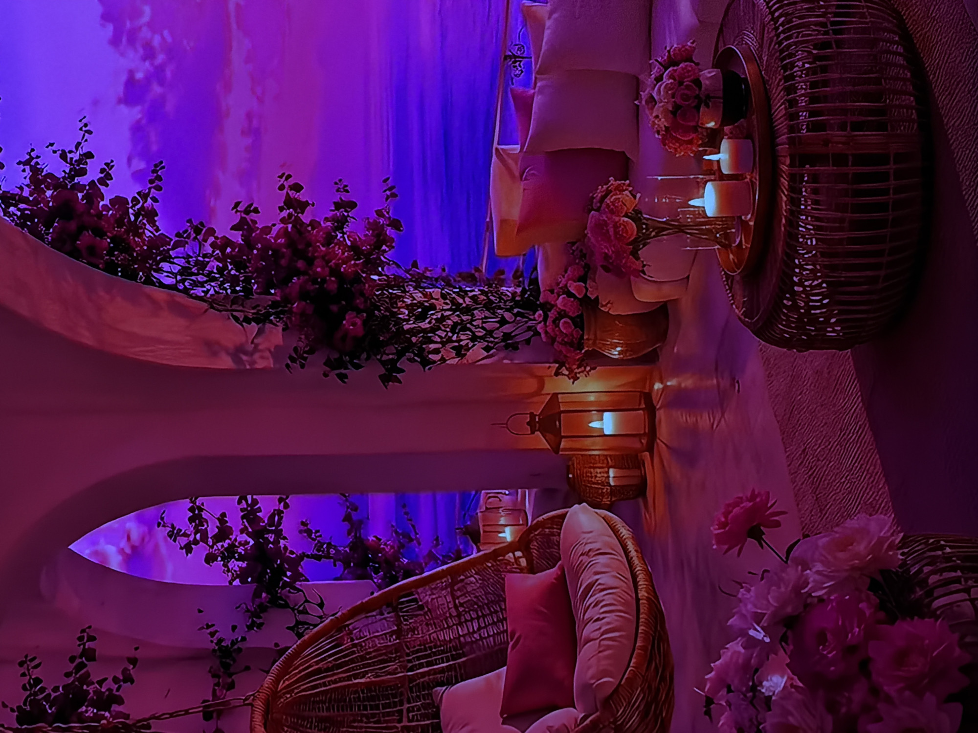 This image captures a serene and romantically lit indoor or covered outdoor space, set in Sofiivska Borshchahivka, Ukraine, during the evening or night. The scene is dominated by soft purple and blue ambient lighting, creating a tranquil atmosphere.
The primary subjects include lush green foliage densely covered with small red or pink flowers, which cascades over light-colored architectural features, possibly archways or decorative elements. A prominent wicker hanging chair, furnished with cream and pink cushions, occupies the foreground, suggesting a comfortable seating area.
Throughout the space, various decorative elements enhance the ambiance. Multiple lanterns with glowing candles emit a warm, soft light that contrasts with the cool purple and blue background illumination. Vases filled with bouquets of pink roses or peonies are visible, adding to the romantic decor. Another wicker basket or tray, also holding glowing candles, is present on a surface. The overall color palette is rich in purples, blues, and pinks, with warm accents from the candle light and natural wicker.
No people are visible, and no specific activity is depicted, implying a static scene set for relaxation, contemplation, or a quiet gathering. No text is visible in the image.
