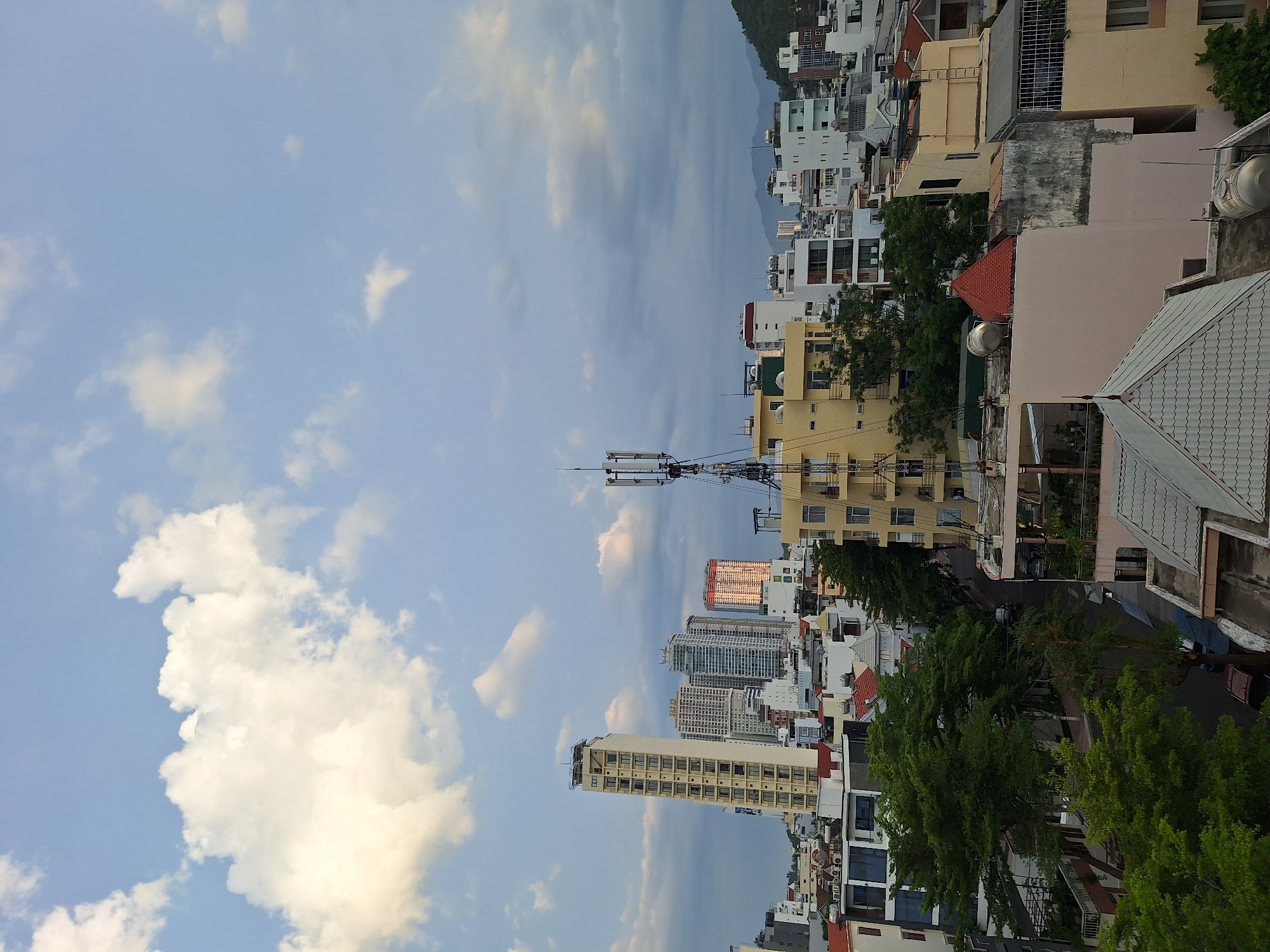The image captures an urban cityscape in Nha Trang, Vietnam, under a partly cloudy sky. In the foreground, various buildings with tiled roofs and balconies dominate the scene. A tall, slender, cream-colored building with many windows stands prominently to the left, characteristic of modern high-rise architecture. Other buildings of varying heights and architectural styles, including some with orange and white facades, are clustered together. A prominent cellular tower with multiple antennas is positioned to the right of center. Dense green trees are interspersed among the buildings, suggesting a well-established urban area with green spaces. In the background, more buildings stretch towards rolling hills covered in vegetation. The sky is a pale blue with large, fluffy white clouds, and some darker, more diffuse clouds suggesting possible weather changes. The overall impression is of a bustling city on a partly overcast day. There are no people visible in the image.