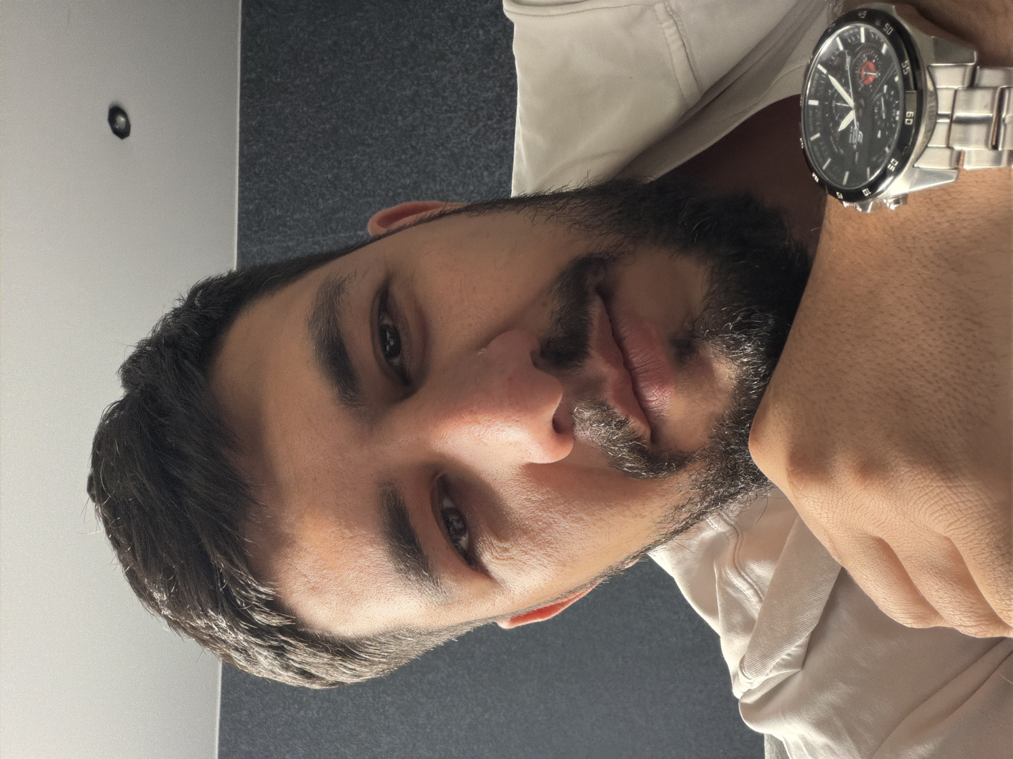 A close-up shot focuses on a man's face and wrist. He has dark hair and a beard, with light skin. He is wearing a light-colored shirt. A metal wristwatch with a dark face is prominent on his wrist. The background is indistinct, appearing to be a gray wall with some white paneling. The location is not discernible from the image.