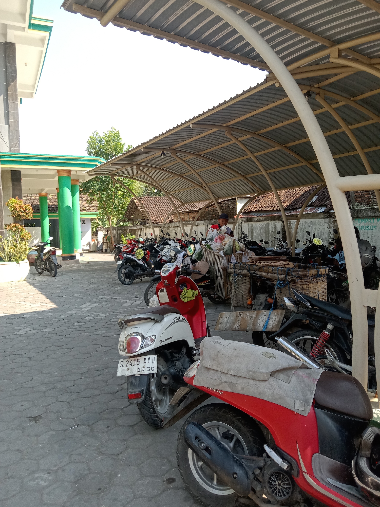 The image depicts a parking area under a corrugated metal roof supported by a white, curved metal frame, likely a covered motorcycle parking lot. The ground is paved with interlocking gray stones. In the foreground, two scooters are parked: a red and black scooter with a light gray cloth draped over its seat, and a white scooter with a license plate reading "S 2415 AAV 11-26". Behind these, a line of parked motorcycles, mostly red and black, extends further back. To the left, a building with green pillars and accents is visible, with potted plants near its entrance. Further back, traditional Indonesian houses with tiled roofs and a brick wall can be seen. A person, wearing a red shirt and a dark cap, stands behind a cart filled with goods, possibly a vendor. The scene is outdoors, under bright sunlight, casting shadows on the ground. The overall impression is a typical scene in a town in Indonesia, likely a public or communal space where people park their vehicles.