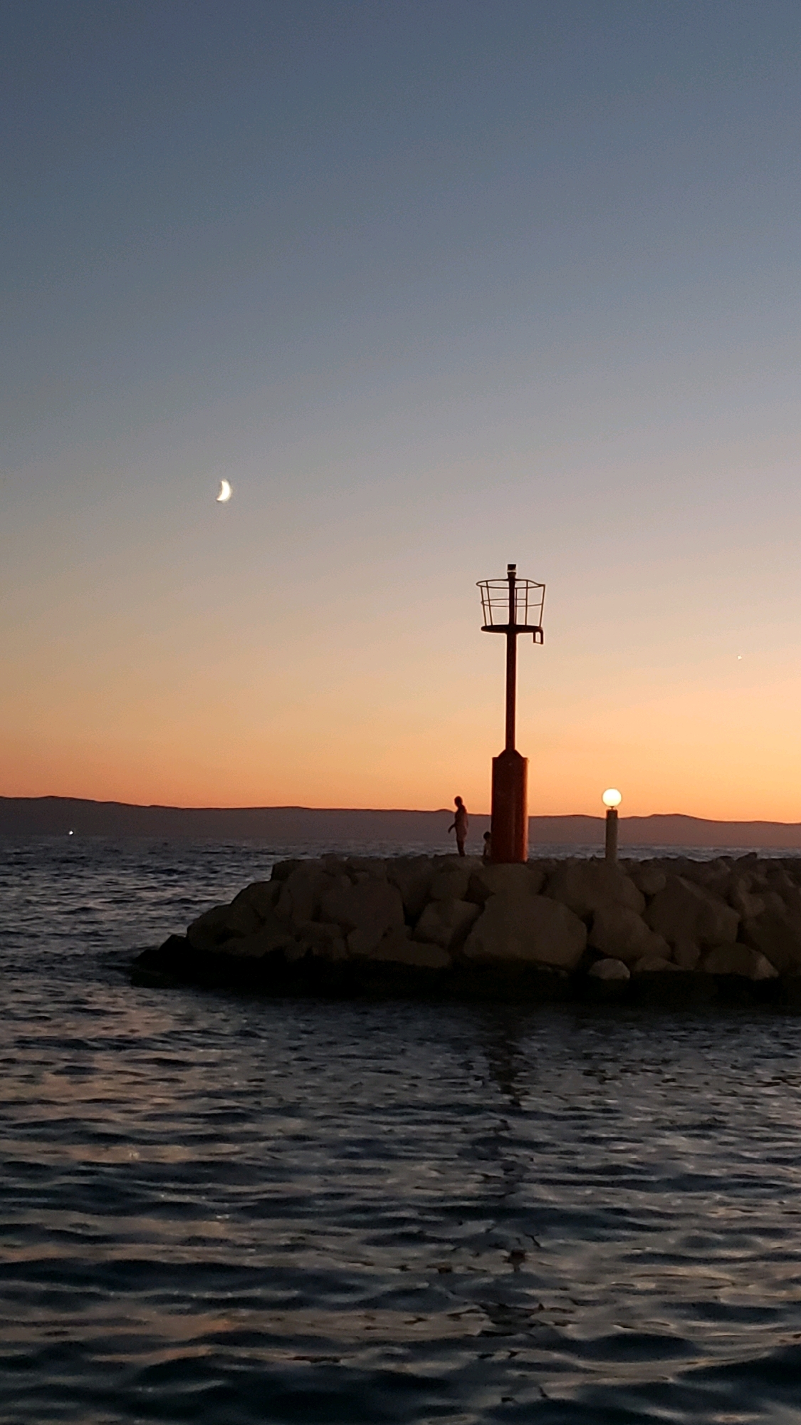 This image depicts a twilight scene on a rocky breakwater, likely in Općina Tučepi, Croatia. A red navigational lighthouse with a cage-like structure at its top stands prominently on the breakwater. Next to it, a smaller, circular light fixture glows. Two human figures, silhouetted against the darkening sky, stand on the breakwater near the lighthouse. The sea, with gentle ripples reflecting the sunset colors, occupies the lower portion of the frame. In the distance, a hazy, mountainous coastline is visible. The sky is a gradient of soft blues and oranges, with a sliver of the crescent moon visible in the upper left quadrant. The overall impression is one of tranquility and the end of a day by the sea.