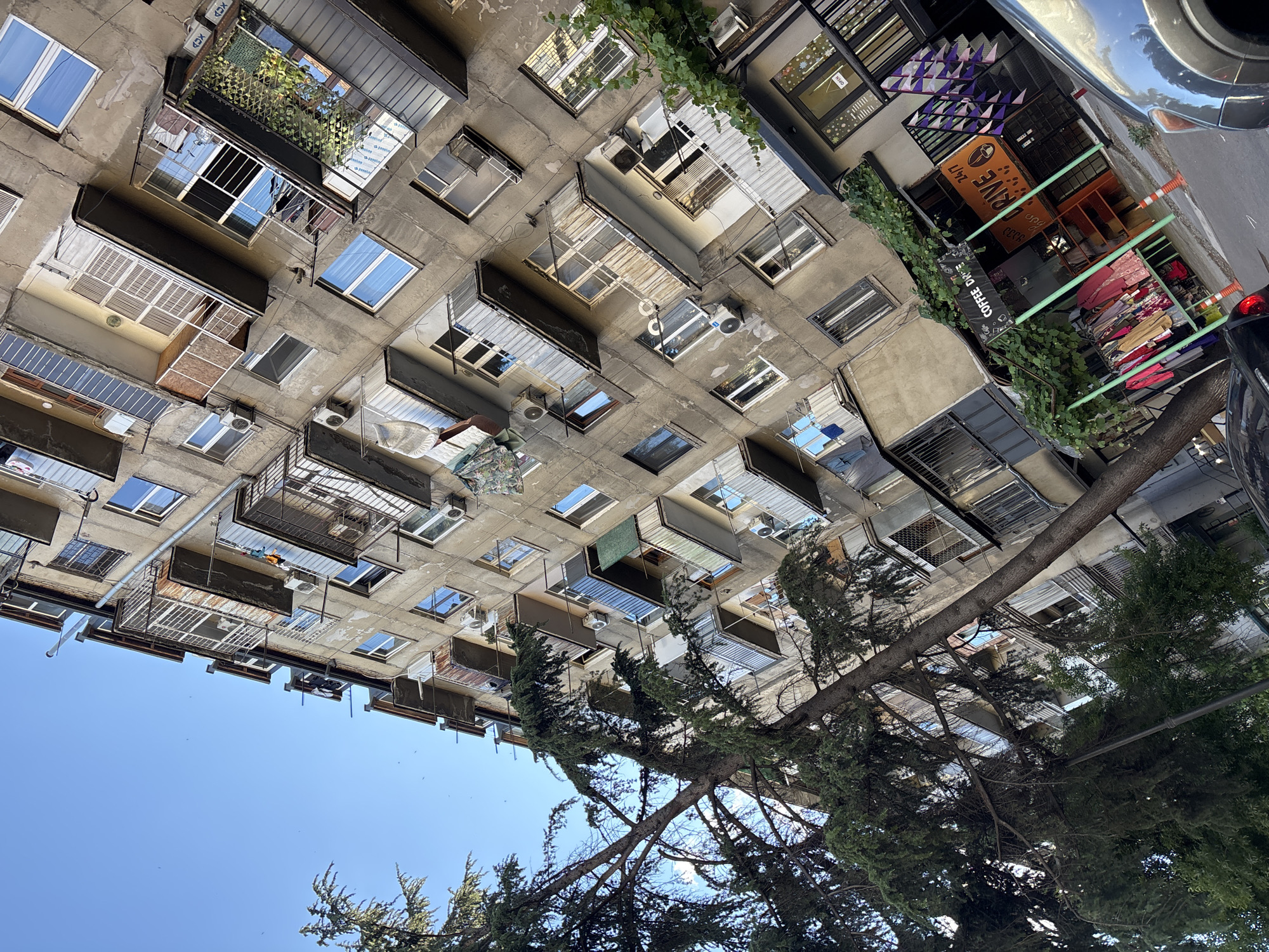 The image shows an apartment building with a row of balconies on multiple levels, with some of the balconies enclosed. A large tree with green foliage is positioned to the left of the building, partially obscuring it. At the base of the building, there is a small establishment with green supports and a sign that says "COFFEE DRIVE 24/7". Clothing is displayed on a rack in front of this establishment. Orange traffic cones are placed on the sidewalk. A dark-colored car is parked in the foreground. The sky is clear blue. The image is taken at an upward angle.