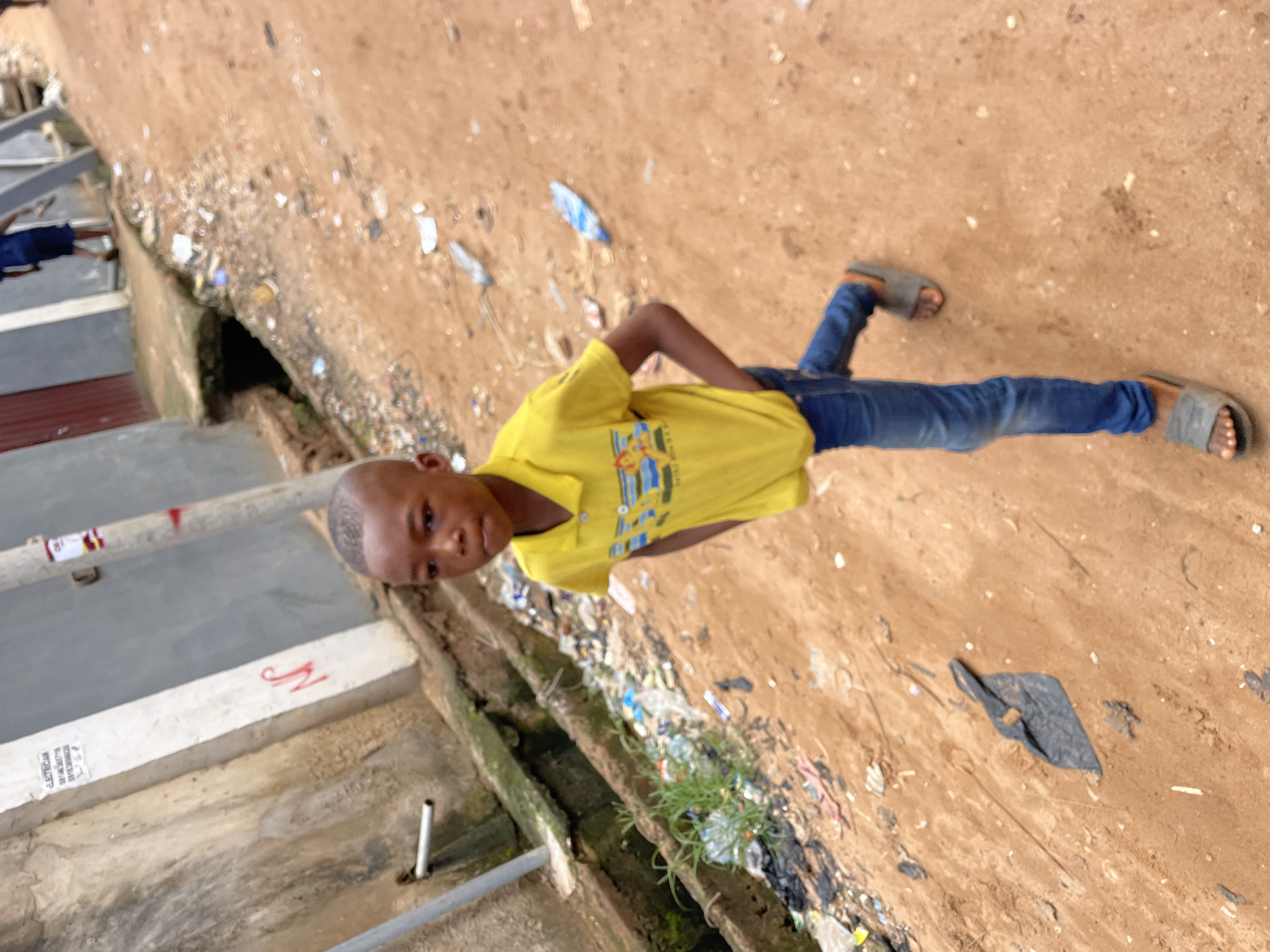 A young boy with closely cropped hair stands on a dirt path alongside a concrete structure, possibly a covered drainage system, in what appears to be a marginalized area. The boy wears a yellow short-sleeved shirt and blue jeans, with sandals on his feet. The ground around him is littered with trash. The scene suggests a low-income neighborhood in Onitsha, Nigeria. The concrete structure runs parallel to the dirt path, and the entire scene suggests a lack of proper waste management and infrastructure. A building is visible on the left side of the frame.