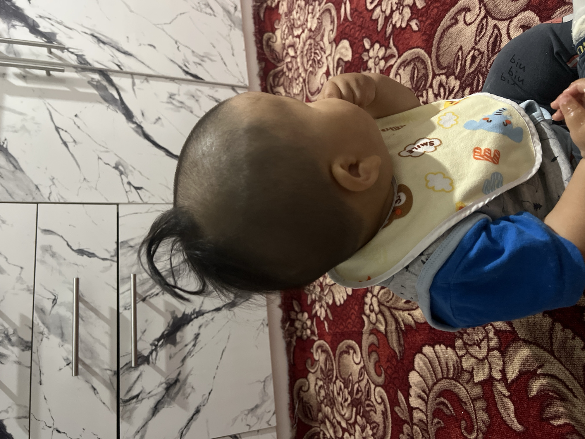 In an indoor setting, a young child, seen from behind, sits on a red patterned carpet. The child wears a blue and gray outfit with a yellow bib featuring cartoon designs. The child's head is partially shaved with a small tuft of hair at the crown. In the background, a white cabinet with marble-patterned doors is visible. The scene is brightly lit.