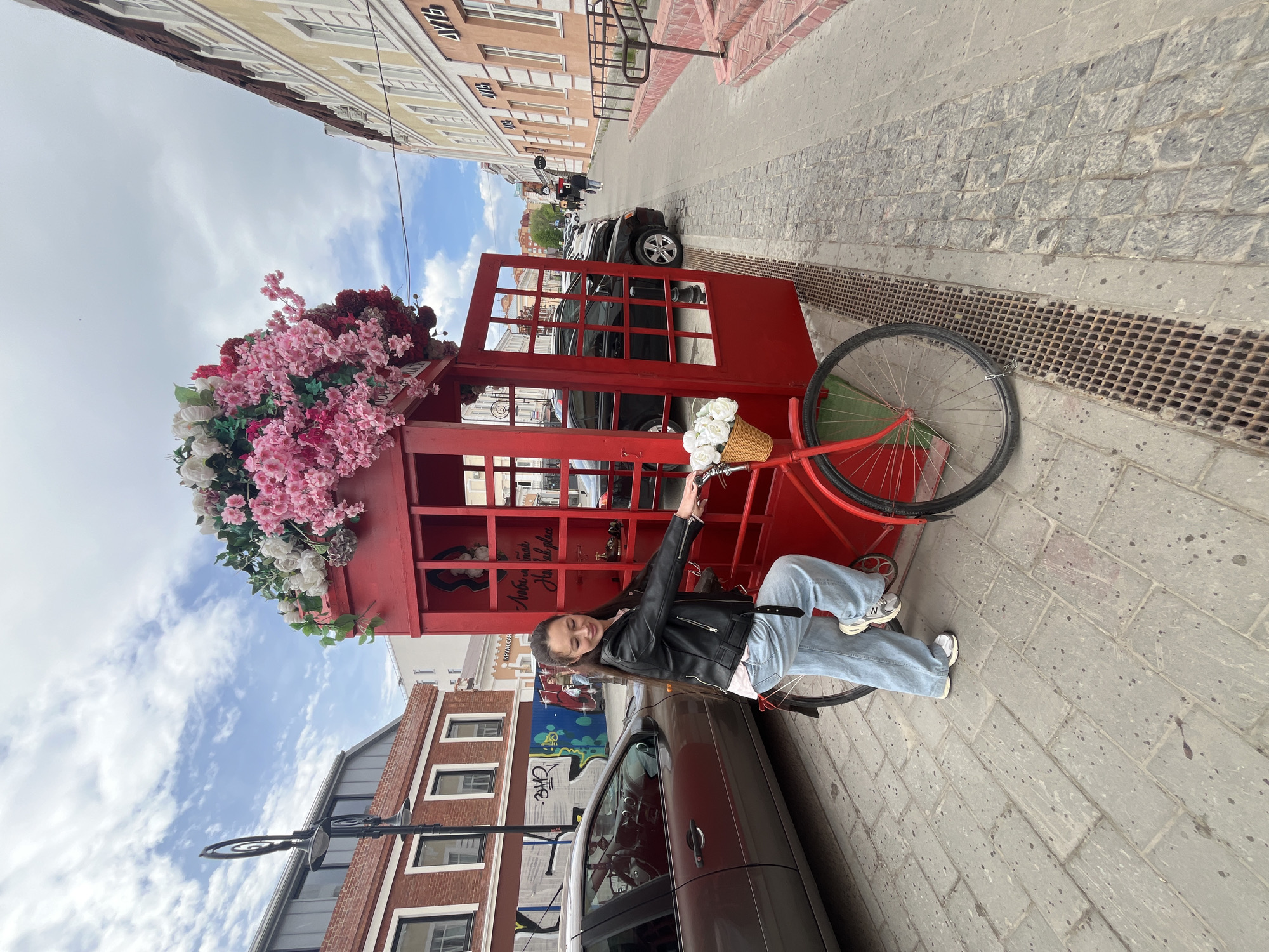 A young woman is sitting on a red bicycle next to a red telephone box decorated with flowers. The scene is located on a city street in Ulyanovsk, Russia, as identified by Cyrillic text on nearby buildings. The woman is wearing a black leather jacket, light blue jeans, and white sneakers. She is holding the handlebars of the bicycle. The telephone box is filled with various decorative items. Cars are parked along the street, and other pedestrians are visible in the background. The sky is overcast with scattered clouds. The street is paved with cobblestones and features a drainage grate along the edge.