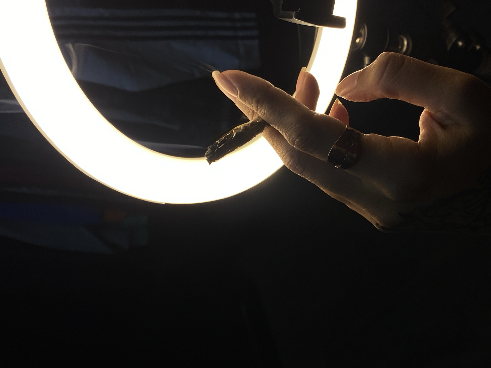 A close-up shot focuses on a person's hand holding a partially smoked, dark-colored cigarette or joint. The hand has a gold ring on one of the fingers and a tattoo visible on the wrist. The background features a circular light ring that provides illumination, with the rest of the scene mostly dark. The setting suggests an indoor environment with a modern light source. There is no evidence that places the scene in Llavallol, Argentina.