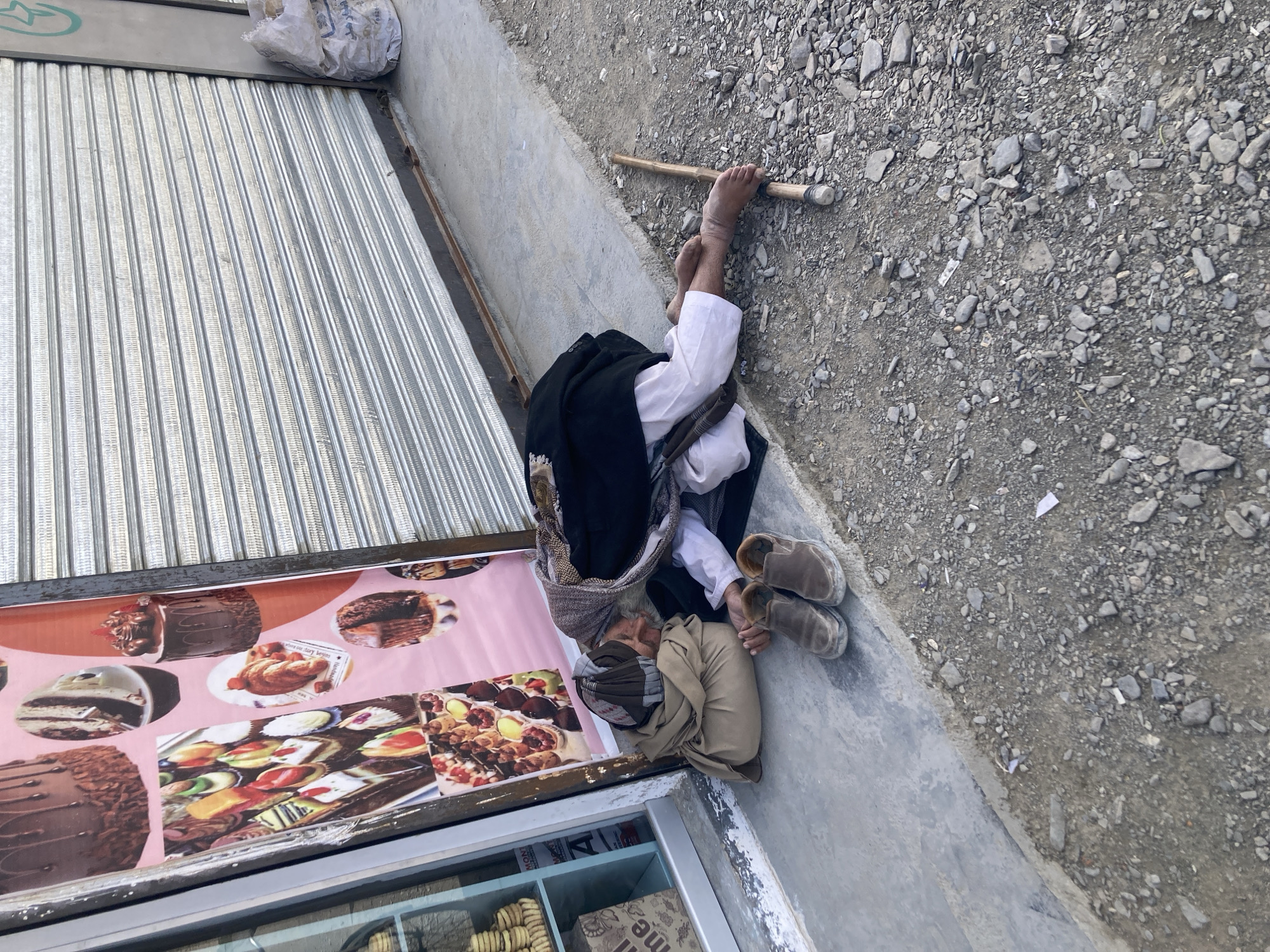 An elderly man is sleeping on the sidewalk in front of a bakery in Gulistan, Pakistan. He is curled up with his head resting on his folded arms, and his feet are bare, resting on a walking stick. His shoes are placed neatly next to him. He wears a turban, a light-colored tunic, and a dark vest. The bakery's storefront is visible with a closed metal shutter and a display case featuring images of cakes and pastries. The sidewalk is concrete, and the ground next to it is covered with gravel and small rocks.