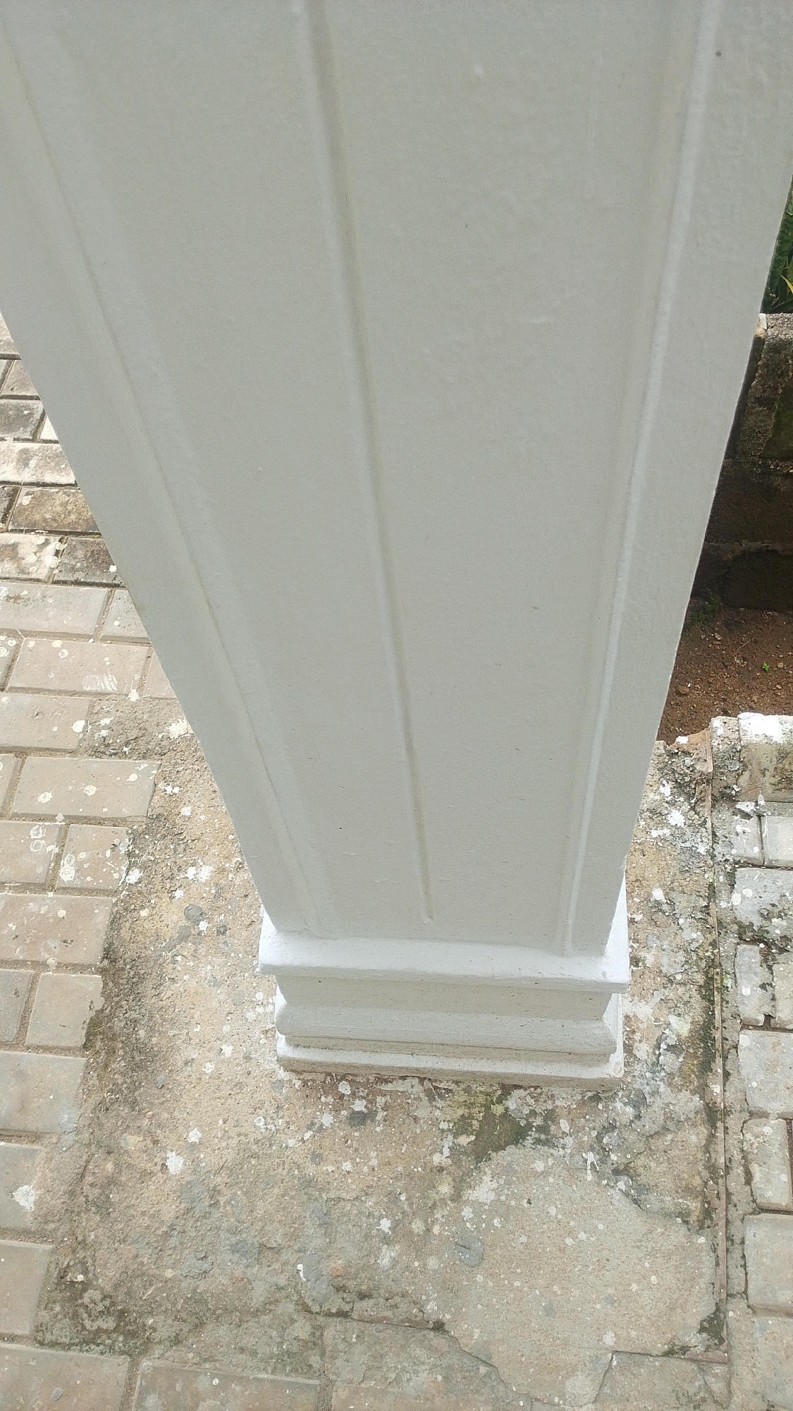 The image shows a close-up, low-angle view of a white, fluted architectural pillar standing on a paved outdoor surface. The pillar has a layered base with horizontal grooves. The paving consists of light brown rectangular tiles interspersed with patches of concrete or dirt. Some white splatters, possibly from birds, are visible on the ground and the lower part of the pillar. To the right of the pillar, a darker area of soil and some green foliage can be seen, suggesting a garden or natural environment. The lighting suggests it is daytime, and the overall scene appears to be an exterior architectural setting, possibly in a residential or public space in Balewa, Nigeria, given the location context. There is no text visible in the image.