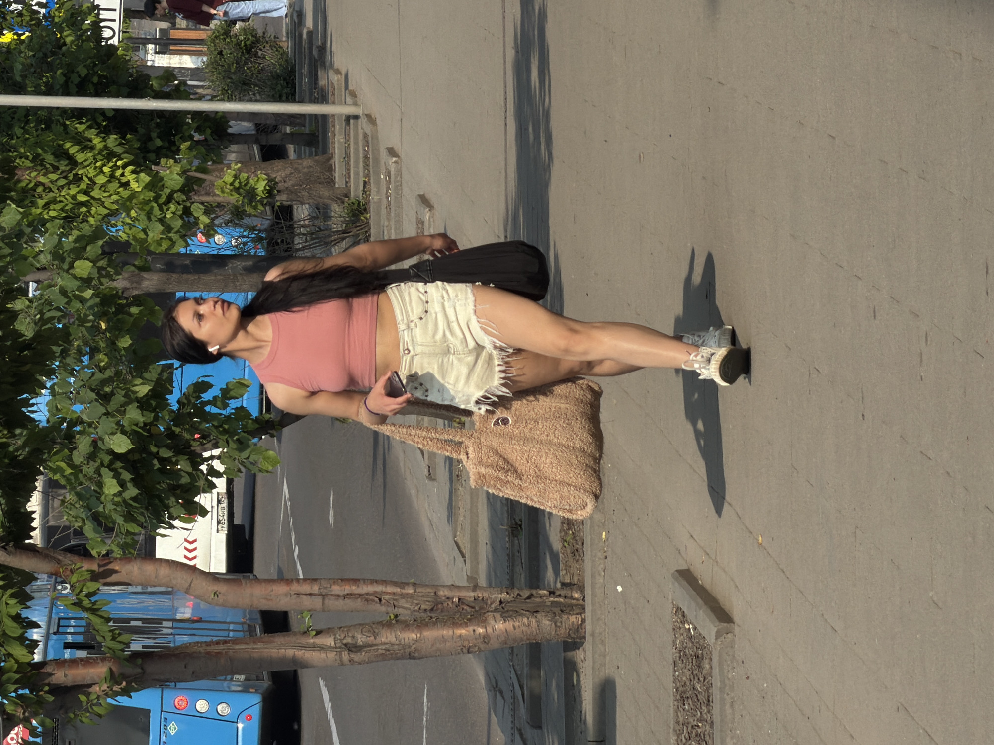 A woman walks on a sidewalk in Krasnoyarsk, Russia, on a sunny day. She is wearing a pink crop top and distressed denim shorts, carrying a beige textured bag on her left arm and a black bag on her right shoulder. The woman has dark hair pulled back and appears to be wearing white earbuds. She is looking to her right. In the background, a blue bus is visible, along with trees and a street lamp. The sidewalk is paved with concrete slabs, and there is a curb and some vegetation bordering the walkway. The overall scene depicts an urban environment with a focus on a pedestrian.
