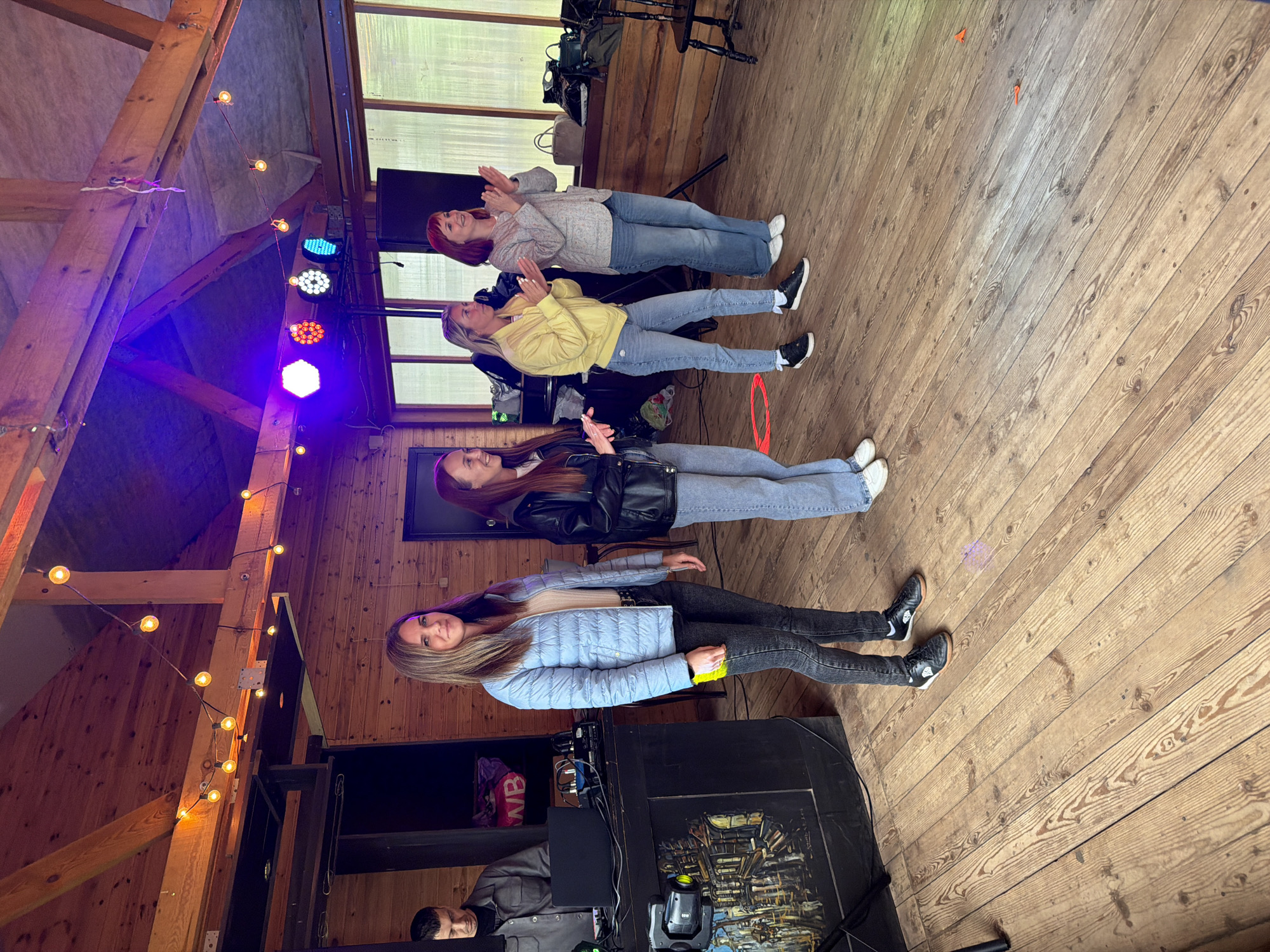 Four women are standing in a line on a wooden floor, inside a room with a timber-framed ceiling decorated with string lights and stage lights. The walls appear to be wood paneled. The women are dressed in casual clothing, including jeans, jackets, and sweaters. The topmost woman has red hair. All four are looking up and have their hands in a clapping position. There is a person visible near the bottom left, next to a sound system. The lighting suggests it could be a party or event. The location of the city of Chaikovsky, Russia is not discernable from the image.