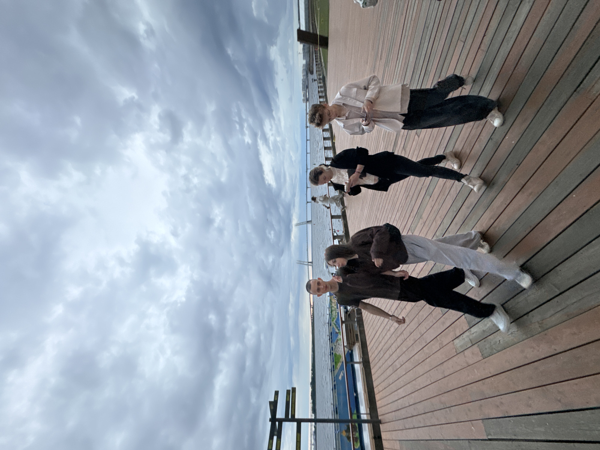 Four young people, appearing to be teenagers or young adults, are walking on a wooden boardwalk by a body of water under a cloudy sky. On the left, a male with short hair wears a dark t-shirt and black pants. Beside him, a female wears a dark hooded sweatshirt and light grey sweatpants. Further to the right, a female in a white top and black jacket holds a smartphone, and beside her, a male wearing glasses, a light beige jacket over a light-colored shirt, and dark pants also looks at his phone. In the background, a bridge spans the water, and buildings are visible in the distance. The boardwalk has visible planks and appears to be part of a waterfront promenade. The scene is likely located in St. Petersburg, Russia, given the architectural style of the bridge and the presence of a large body of water consistent with the Neva River or the Gulf of Finland. The lighting suggests it is late afternoon or early evening.