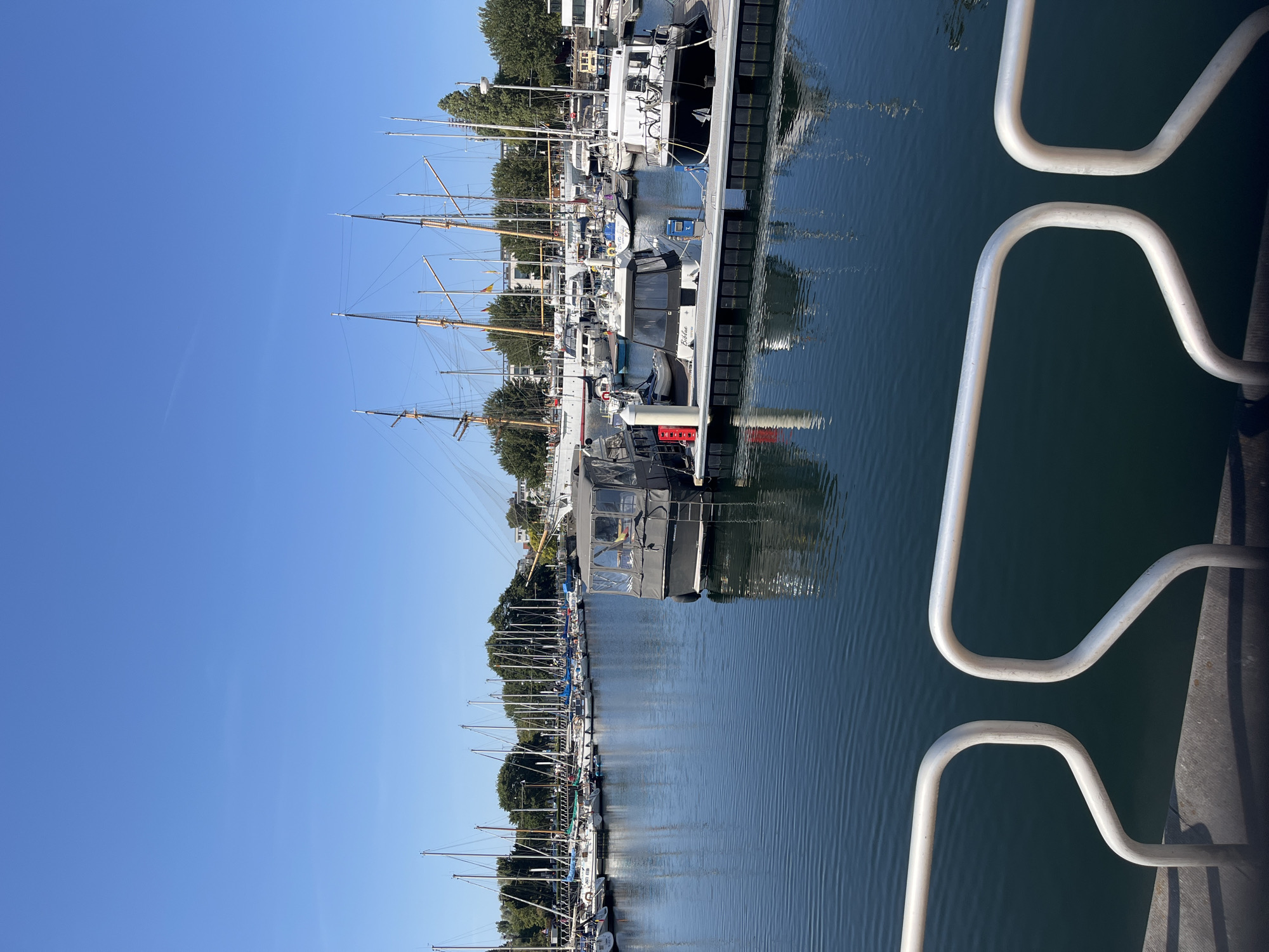 The image depicts a marina in Ostend, Belgium, on a clear, sunny day. The foreground features the edge of a dock with white metal railings. The water is a deep blue-green, reflecting the sky and the boats. A variety of boats are moored in the marina, including sailboats with tall masts, motorboats, and a larger ship with multiple masts visible in the background. One motorboat in the foreground is covered with a gray canvas. Trees and buildings line the background of the marina. The scene is calm and orderly, indicative of a peaceful day at the harbor.