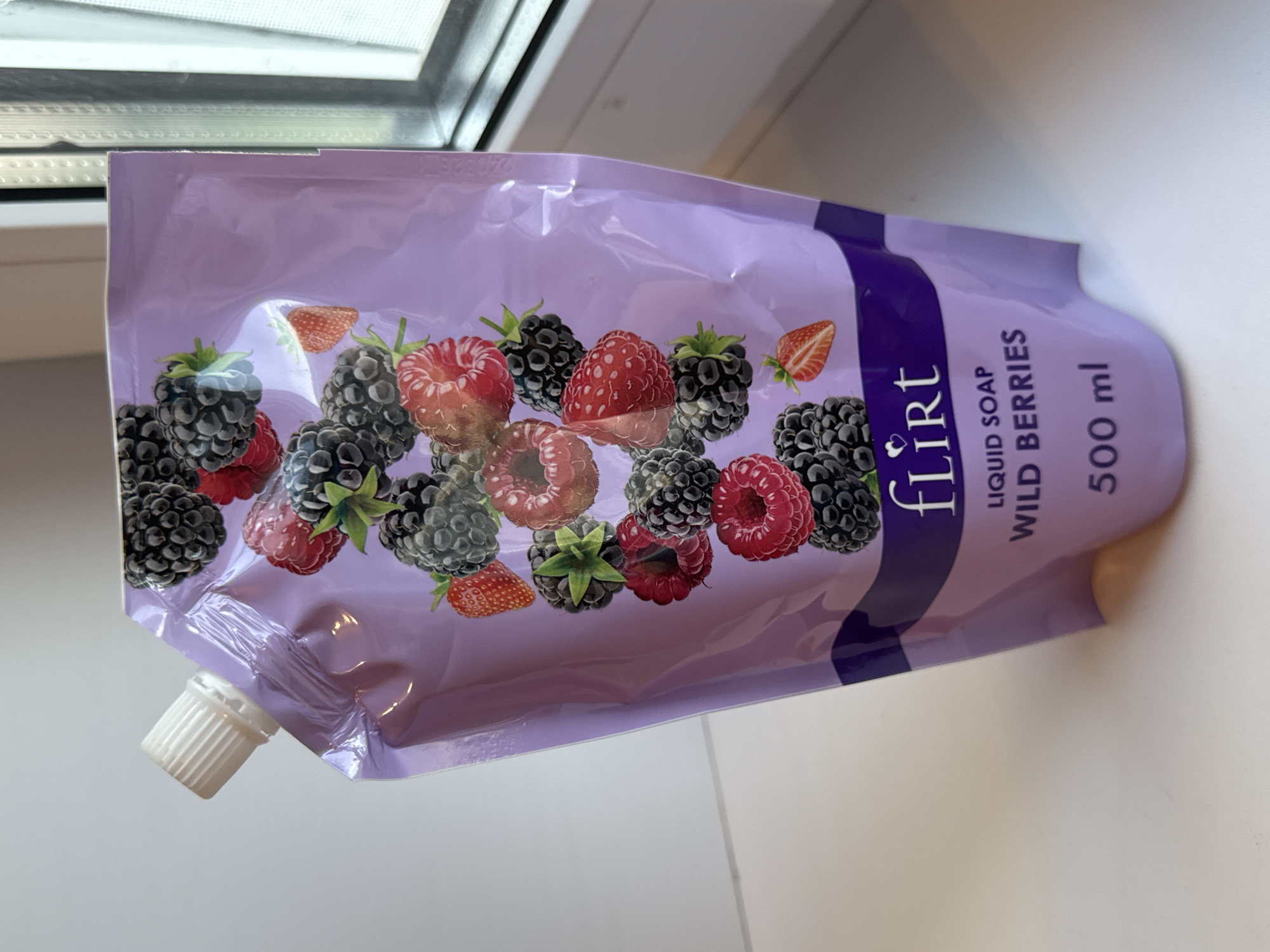 A pouch of liquid soap with a wild berry scent is shown. The pouch is lavender in color and features an illustration of various berries, including raspberries and blackberries. The brand name "FLIRT" is printed in white text, followed by "LIQUID SOAP WILD BERRIES" and "500 ml". The pouch has a white screw-top cap and is positioned on a white surface, possibly a windowsill or table, with a window visible in the background. The lighting suggests it is daytime. The scene is indoors.