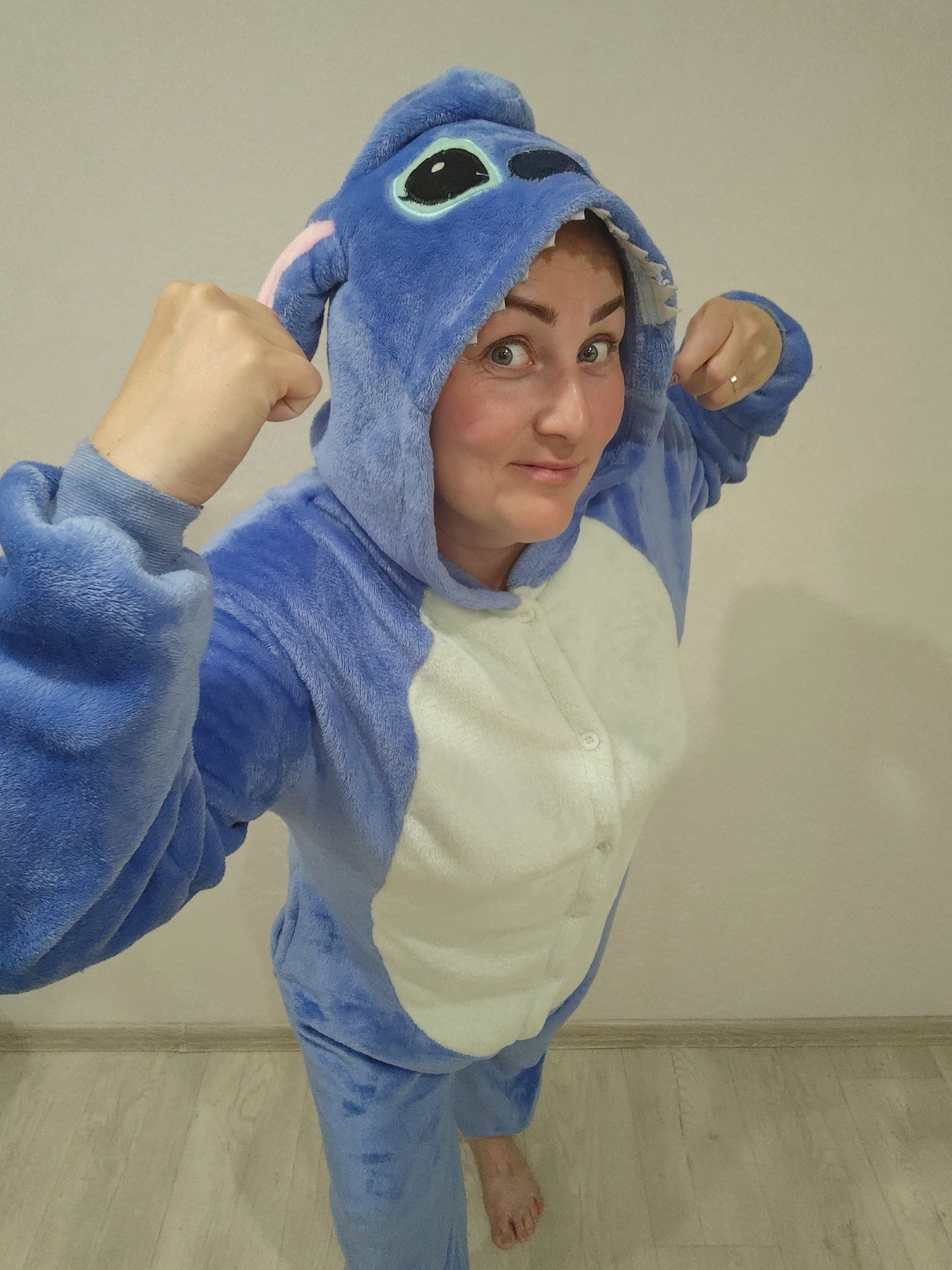 An adult female is depicted standing indoors in the city of Markovskii, Russia. She is wearing a blue and white plush kigurumi, designed to resemble the character Stitch, with the hooded headpiece featuring large black and teal eyes and pink inner ear detailing worn over her head. Her face is fully visible, displaying a direct gaze towards the camera with wide eyes and a slight smile. Her hair is not visible. Both of her hands are clenched into fists and raised to shoulder height. A thin, metallic ring is visible on the ring finger of her right hand. She is barefoot, with her right foot partially visible on the light-colored wooden floor. The background consists of a plain, light beige wall. A narrow, dark baseboard runs along the bottom of the wall where it meets the floor. The scene is illuminated by what appears to be artificial light, casting soft shadows.