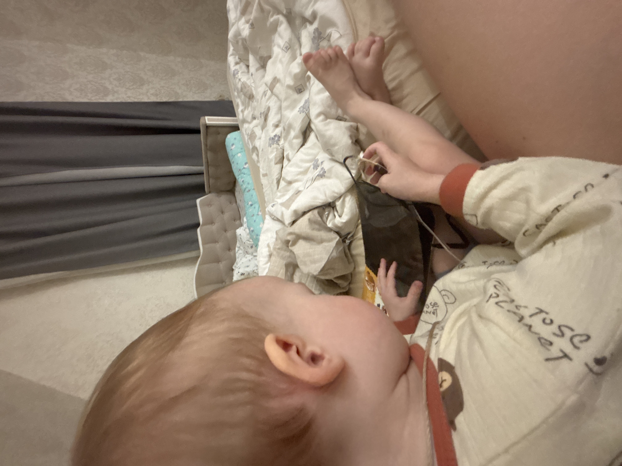 A top-down shot shows a baby in Ферма, Russia, lying on a bed. The baby's head, covered with light brown hair, is at the bottom left of the frame, with the ear visible. The baby wears a light-colored onesie with images, and its arm extends towards the center. The baby's feet are visible at the top, near a pillow covered with a white and blue patterned blanket. A hand, possibly belonging to an adult, is near the baby's feet, holding a black object with a wire. The bed is made with a white patterned comforter. The background includes a gray-toned window and a light-colored wall.