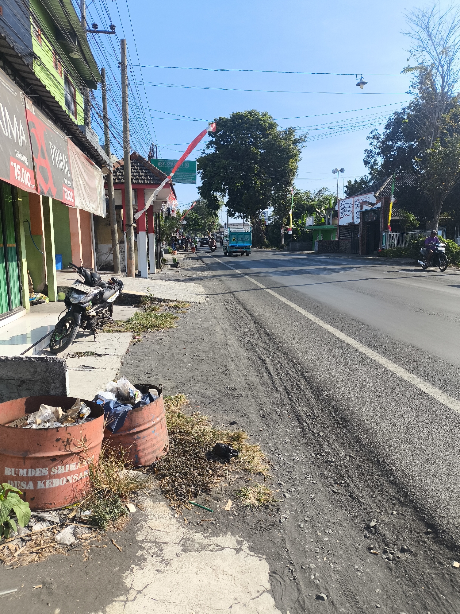 The image depicts a street scene in Labruk Kidul, Indonesia, under a clear blue sky. A paved road runs diagonally through the frame, with a narrow dirt and gravel shoulder on the left. The shoulder is lined with two large, rusty, open-top barrels overflowing with trash. A black motorcycle is parked on a concrete slab in front of a row of shops. These shops have colorful facades, with green and orange being prominent. A large banner with red lettering is visible on the left-most shop.
The road is trafficked by several vehicles, including a light blue truck in the distance and a person on a motorcycle further down the road on the right. The surrounding environment is somewhat urban, with overhead power lines crisscrossing the sky. Trees are present on the right side of the road, and a large, leafy tree is prominent in the middle ground. Red and white Indonesian flags are displayed along the roadside, suggesting a possible local celebration or observance. The overall impression is a functional, everyday street in a populated area.