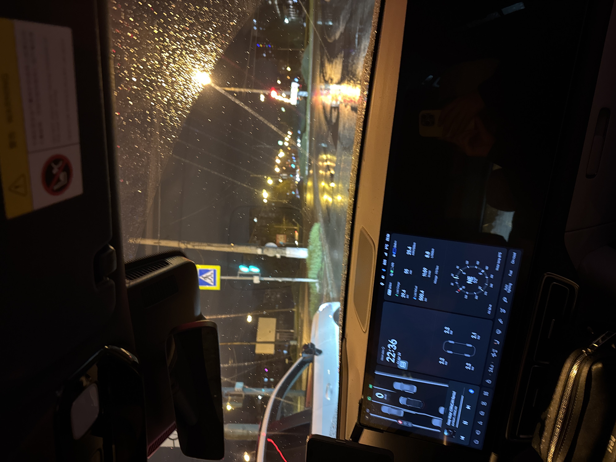 The image captures a nighttime street scene from inside a car, looking out through a rain-splattered windshield. The raindrops refract the bright streetlights, creating a bokeh effect across the view. The car appears to be stopped or parked on a wet road, possibly at an intersection.
On the left side of the frame, part of the car's interior is visible, including a sun visor with a yellow-and-white warning label attached. The label features a red circular symbol with a diagonal line through an icon, likely indicating an airbag warning for child seats. A dashcam or similar device is mounted near the rearview mirror, partially obscured.
Through the wet windshield, the street reveals several details: bright streetlights illuminate the scene, and a traffic light showing a green signal is visible in the distance. To the left, a yellow square road sign with a blue border and a white triangle containing a black pedestrian figure marks a pedestrian crossing. The road surface is visibly wet, reflecting the artificial lights. In the middle ground, the front of a white vehicle is partially visible to the right of the car's viewpoint.
Dominating the right side of the frame inside the car is a large, vertically oriented digital infotainment screen. The screen displays various car metrics and information:
- The time "22:36" and date "10 Mar 24".
- Battery information, including "56%" (likely charge level) and an estimated charging time.
- Energy consumption figures, such as "29.4 kWh" and "500.4 Wh".
- Tire pressure readings: "2.2", "2.4", "2.2", "2.2" (likely bar).
- A compass displaying "NE 67".
- A map-like interface showing the car's position relative to other vehicles.
- A music player interface, showing "Deep House (GRANICAN REMIX)" as the current track.
- Standard status bar icons for 5G network, Wi-Fi, and battery.
A black zippered bag or purse is visible next to the screen on the far right. A reflection of a hand holding a phone with a yellow case can be seen on a dark surface further back in the car, suggesting someone is either taking the photo or using a device. The overall atmosphere is quiet, characteristic of a rainy night.