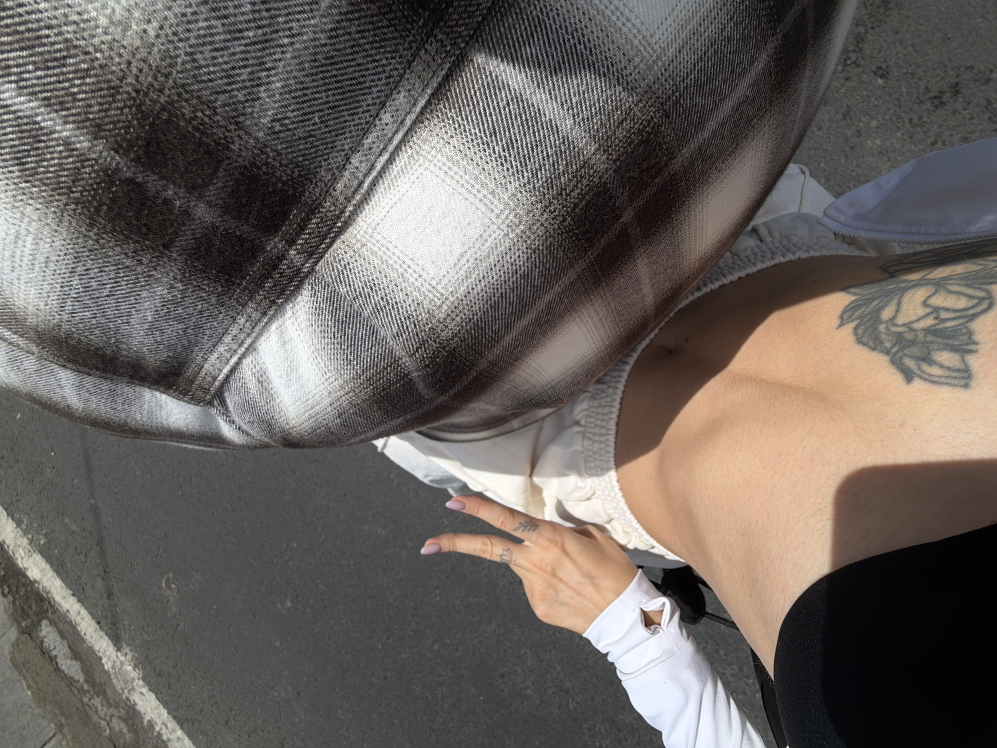 A first-person perspective shot shows a person's midriff, torso, and hand, with a tattoo visible on the lower abdomen and another on the upper thigh. The hand is raised, fingers extended in a peace sign, and a portion of a plaid shirt is visible in the upper right corner. The individual is wearing light-colored shorts and a white long-sleeved top. The scene appears to be outdoors, on asphalt, possibly a street or parking lot, given the presence of a white line marking. The lighting suggests a sunny day. The location is North Kuta, Indonesia.