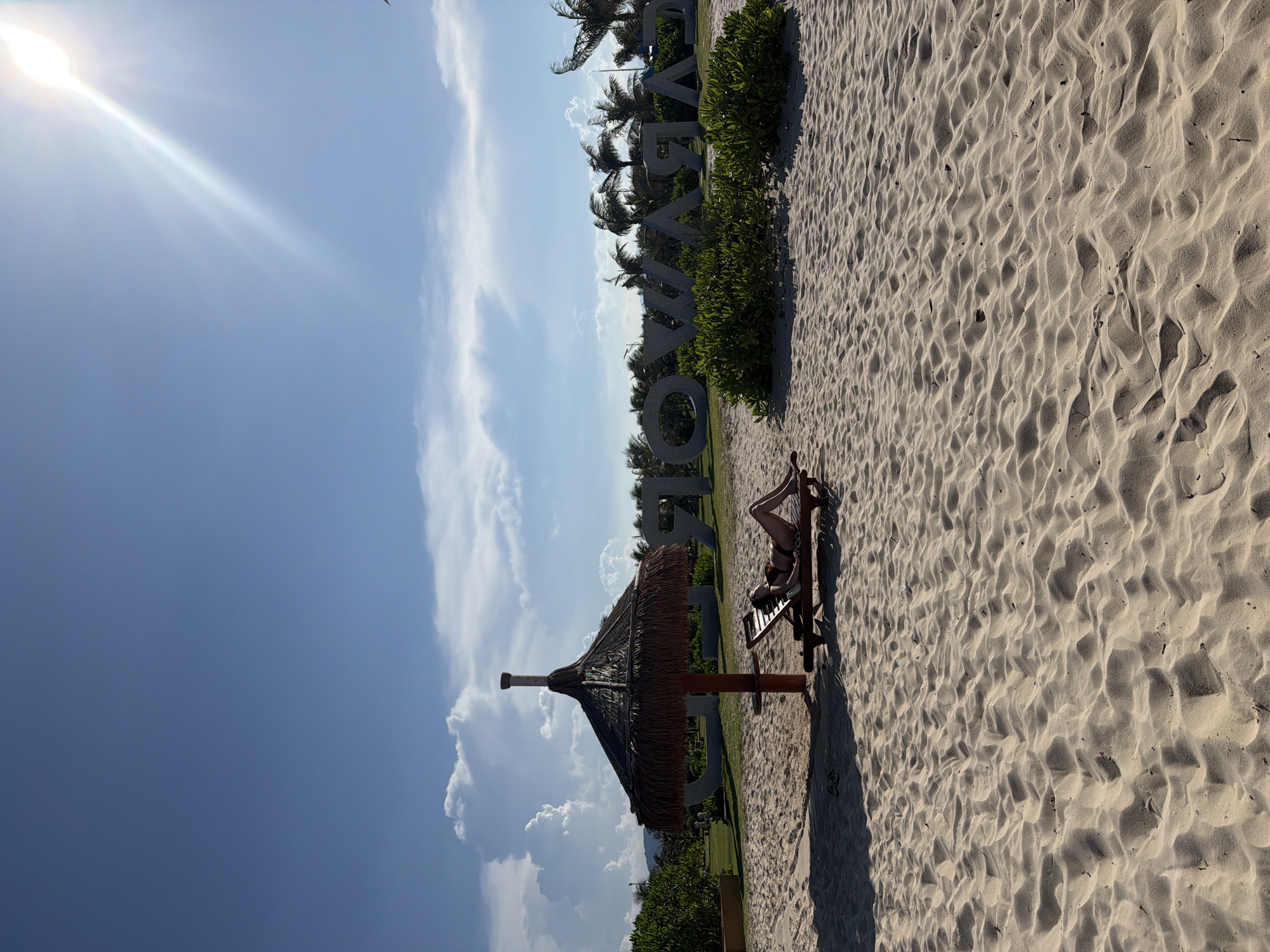 A woman in a bikini reclines on a lounge chair under a thatched umbrella on a sandy beach in Cam Ranh Bay, Vietnam. The sun shines brightly in a clear blue sky with scattered clouds. In the background, large stone letters spell out "NHA TRANG" and palm trees are visible. The sand is textured with ripples from the wind.