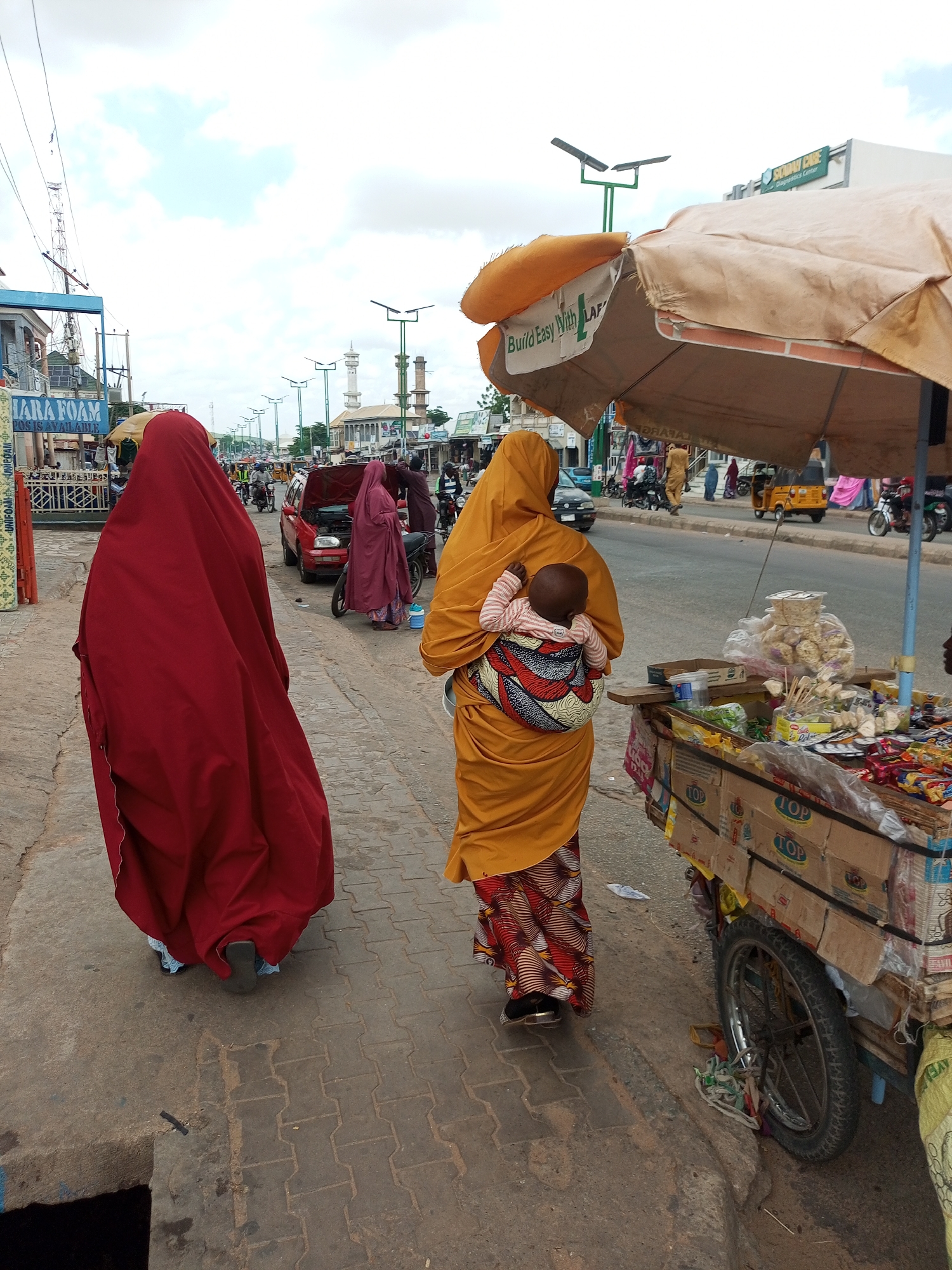 In Arawa, Nigeria, on a cloudy day, two women walk along a paved sidewalk next to a busy street. The woman on the left wears a floor-length, dark red garment that covers her entire body except for her feet. She has on dark, flat shoes and walks with her back to the camera. The woman on the right, also facing away, wears a bright orange hijab and dress, with a patterned skirt in red, yellow, and white. She carries a baby on her back, wrapped in a colorful cloth with geometric patterns of red, white, and blue. Her feet are clad in black shoes. To the right of the women, a cart loaded with various goods, including packaged snacks and other items, is parked by the roadside. The cart is made of wood and cardboard boxes, with a large umbrella providing shade. The street is active with a few vehicles, including a red car with its hood open, a yellow auto-rickshaw, and motorcycles. Buildings line the opposite side of the street, with a mosque's minarets visible in the distance. The environment appears to be a bustling urban commercial area.