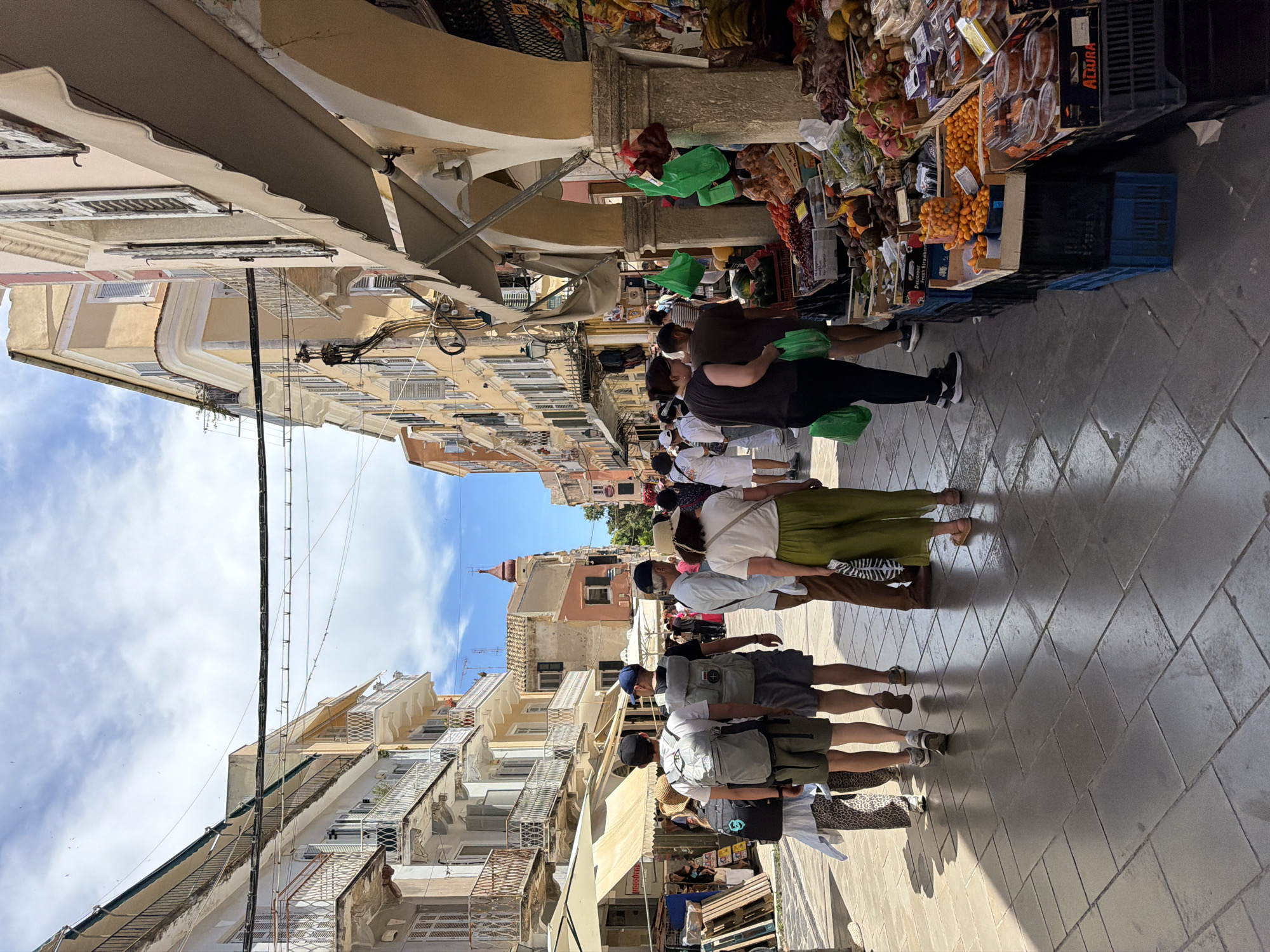 This vibrant image captures a bustling street market scene in what appears to be a historic European town, consistent with Corfu, Greece. The perspective looks down a relatively narrow, paved street lined with multi-story buildings and market stalls.
Numerous people, predominantly adults, are visible walking along the street. Many are seen from behind, suggesting movement deeper into the market. Several individuals are carrying bright green plastic shopping bags, indicating recent purchases.
On the right side of the street, a series of market stalls are set up, overflowing with fresh produce. Various fruits and vegetables are displayed in crates and baskets, including a prominent display of small, round, orange citrus fruits (likely kumquats, for which Corfu is known), alongside other produce. A cardboard box in one stall reads "DELICIOUS," and a blue plastic crate further down has "ALCIRA" printed on it.
The buildings flanking the street feature light-colored facades, traditional wooden shutters on windows, and some balconies. Awnings are extended from some buildings to provide shade over parts of the market. In the distance, a distinctive building with a reddish-orange, spire-like roof stands out against the sky. Overhead, numerous electrical wires and cables crisscross between the buildings.
The street itself is paved with a pattern of grey and lighter-toned rectangular tiles. Strong sunlight illuminates the scene, casting distinct shadows from the people and buildings onto the pavement, indicating a clear, sunny day, likely mid-morning or early afternoon. A blue sky with scattered white clouds is visible between the buildings. The overall atmosphere is lively and typical of a busy local market.