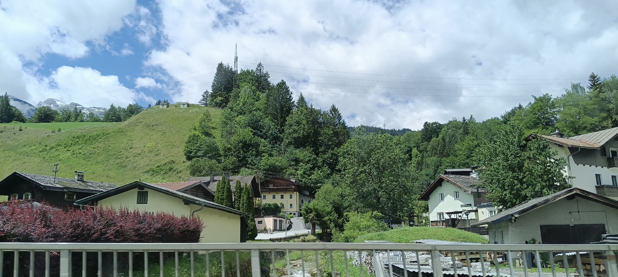The image depicts a village nestled in a valley with a lush green hillside and dense forest. In the foreground, a white metal railing separates the viewer from the scene. Several houses with traditional Alpine architecture are visible, some with light-colored facades and dark roofs, others with darker wood and tiled roofs. A cluster of vibrant red-purple bushes is in front of one of the houses. The hillside behind the houses rises steeply and is covered in grass, with a few scattered trees and two small utility buildings near the crest. A tall telecommunications tower stands atop the hill, surrounded by coniferous trees. Further in the background, a range of majestic snow-capped mountains pierces the sky, partially obscured by clouds. The sky is a mix of blue and white clouds, suggesting a partly cloudy day. The environment is distinctly mountainous and rural.