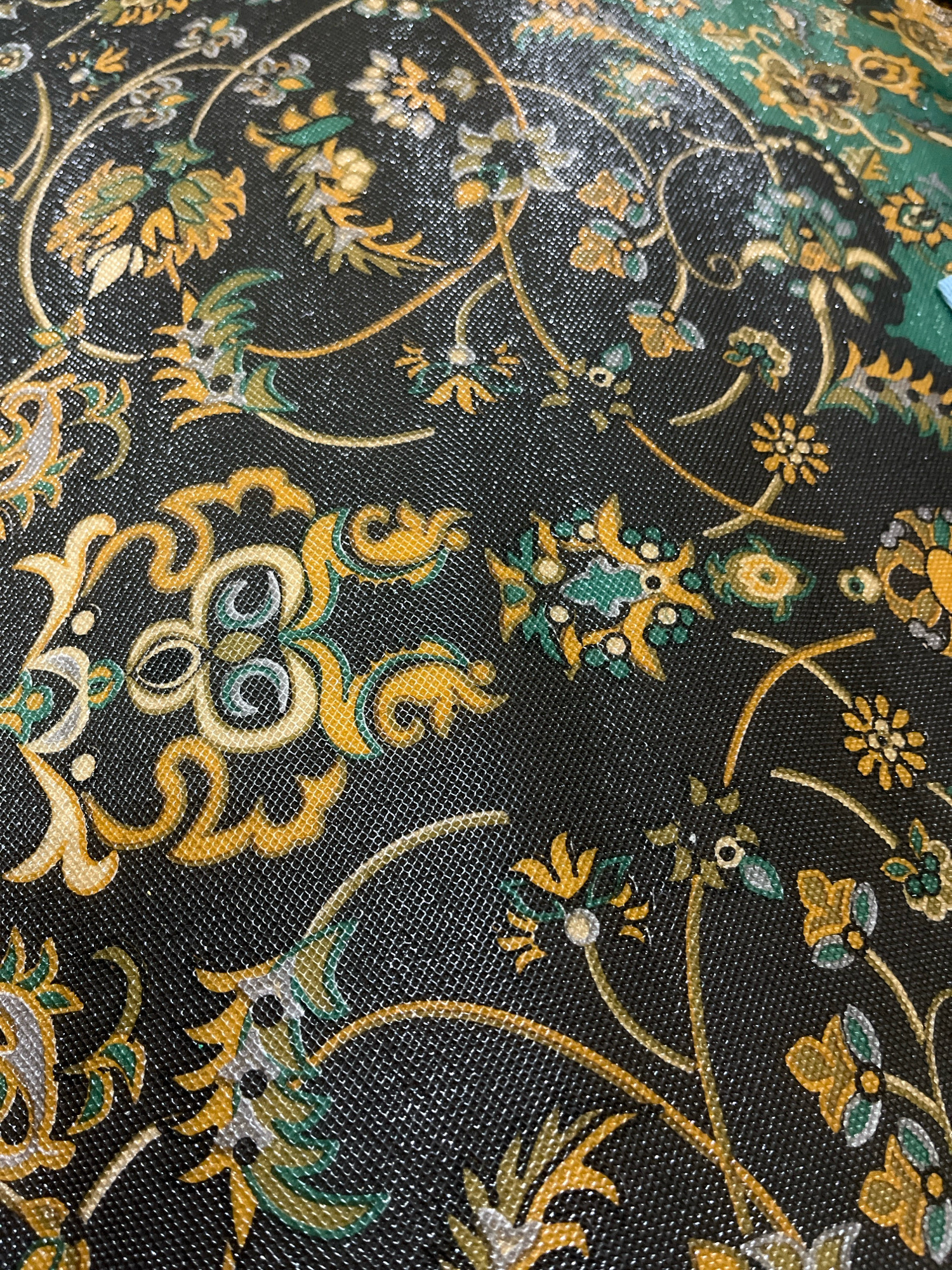The image shows a close-up, angled view of a black fabric with a floral and scrollwork pattern. The pattern is rendered in yellow, green, and white, with some subtle metallic shimmer. The fabric appears to be textured, with a visible mesh-like weave. There are no people, actions, or interactions depicted. The background is the fabric itself. The location in Lambaro Kueh, Indonesia, is not discernible from the visual information provided in the image.