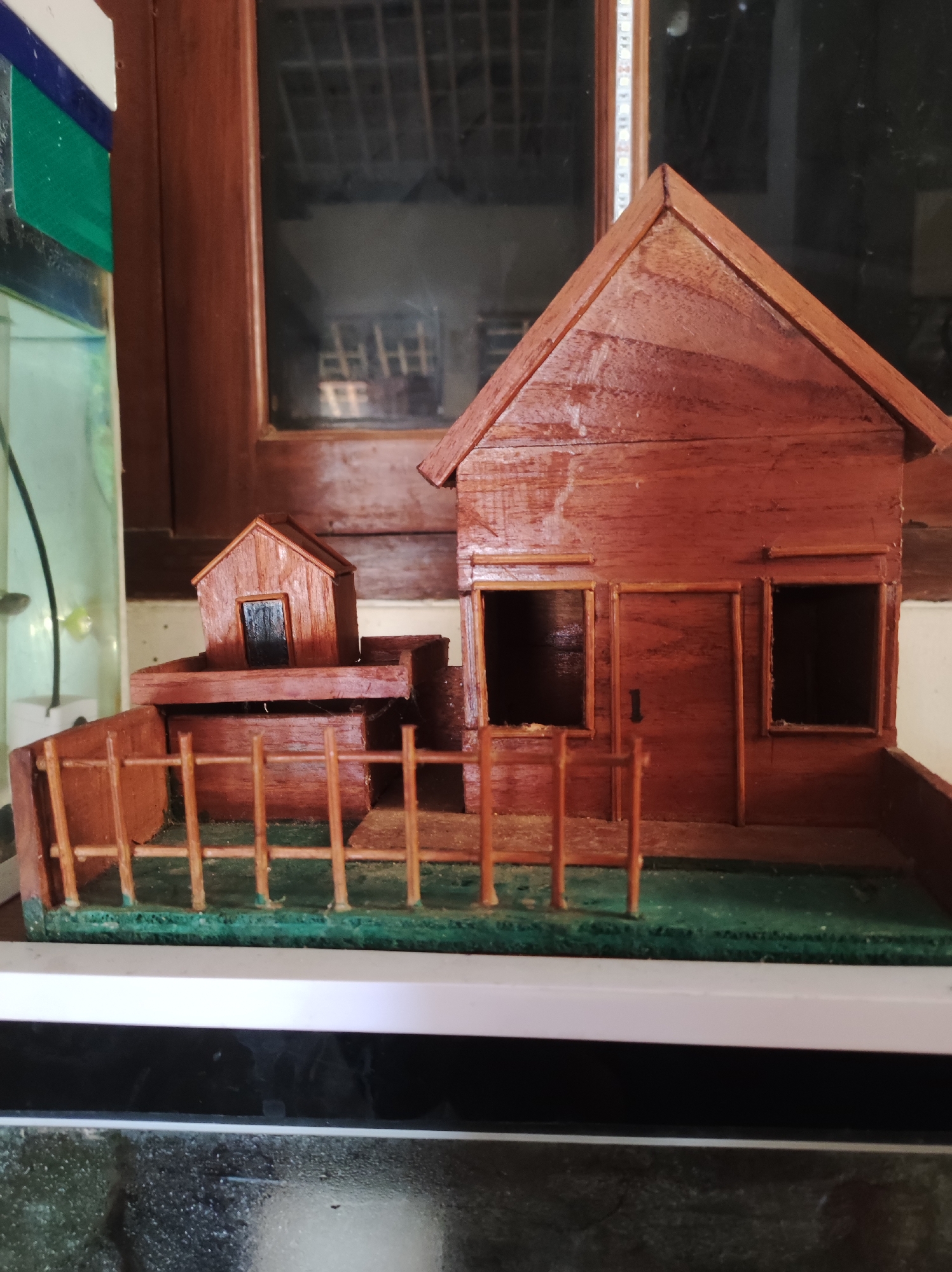 The image shows a close-up of a wooden miniature house with a fence and a small doghouse. The house is made of reddish-brown wood and has two windows and a door on the front. The doghouse is smaller and sits on a raised platform behind the main house. The fence is made of thin wooden posts and rails and encloses a grassy area. The miniature house is set against a dark background, possibly a window or a wall. The overall impression is that of a craft or hobby project. The image does not provide any information about the city of Mojopuro, Indonesia, or any people, actions, or interactions.