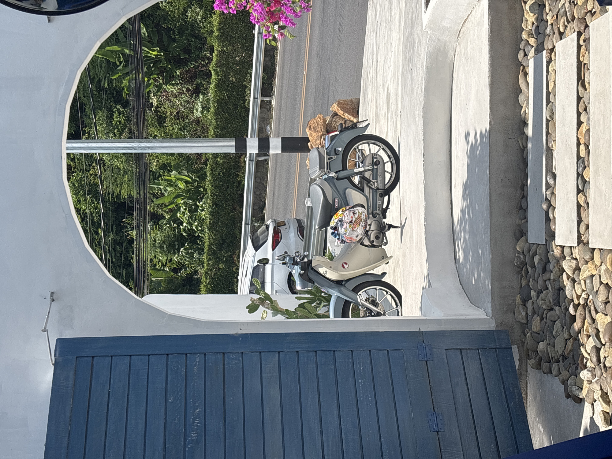 The image, likely taken in Kamala, Thailand, presents an oblique view of an outdoor setting. A blue, vertically-planked gate dominates the lower portion of the frame. Above the gate, a white, semi-circular archway frames the scene. Through the archway, there's a road, a roadside hedge, and a power pole with black and white bands. A scooter is parked partially within the frame of the archway, close to the road. Behind the scooter is a parked white car. To the right of the scooter, outside the arch, is a retaining wall of rounded rocks and what appear to be concrete slabs. A flowering plant with pink blossoms is visible above the road.