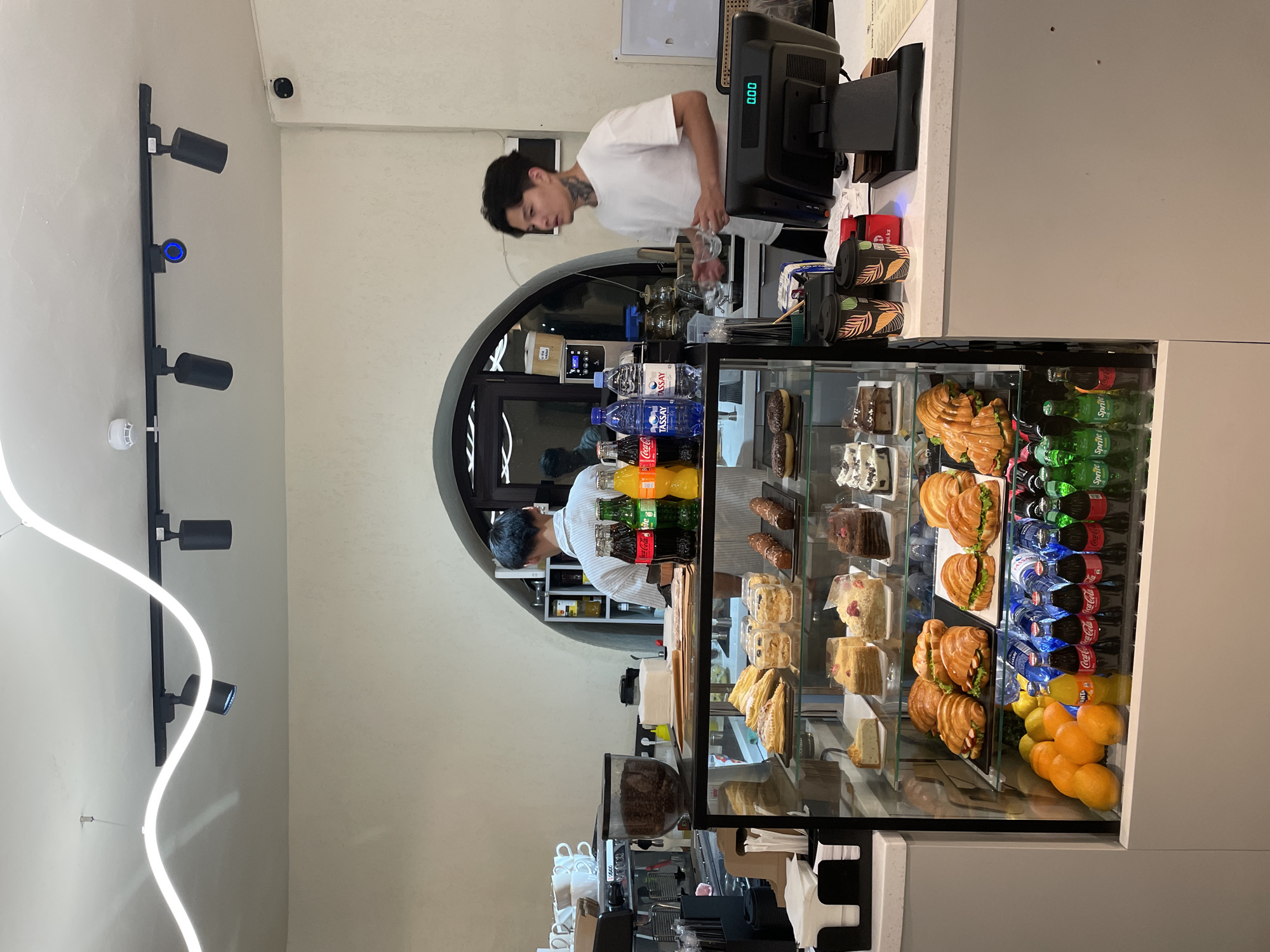 This image shows the interior of a cafe, likely in Almaty, Kazakhstan, given the Coca-Cola and Sprite bottles, as well as Tassay water bottles which are common in the region. The cafe features a display counter filled with various pastries, cakes, sandwiches, and fruits. Behind the counter, two baristas are visible; one is preparing drinks at an espresso machine, and the other is attending to customers. The cafe has a modern aesthetic with a wavy neon light fixture and track lighting on the ceiling. The overall ambiance suggests a casual, contemporary establishment.