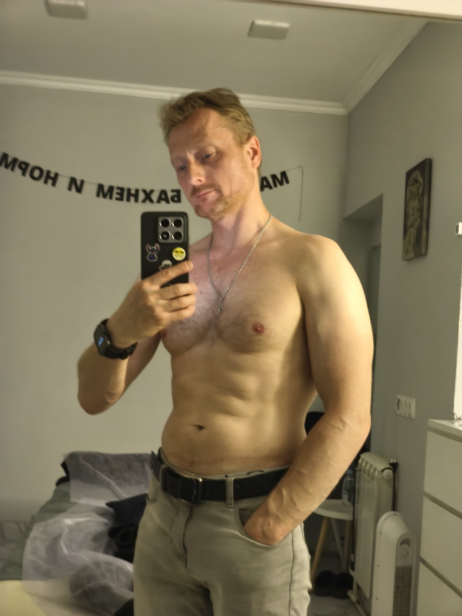 A man with fair skin and light brown hair stands in front of a mirror, taking a selfie. He is shirtless, wearing light-colored pants and a black belt. The room appears to be a bedroom, with a bed visible in the background. A decorative banner with black letters is hung on the wall behind him, and a piece of art is also visible on the wall. A radiator and a white cabinet are present in the room. There are no indications of the location being in Yerevan, Armenia.