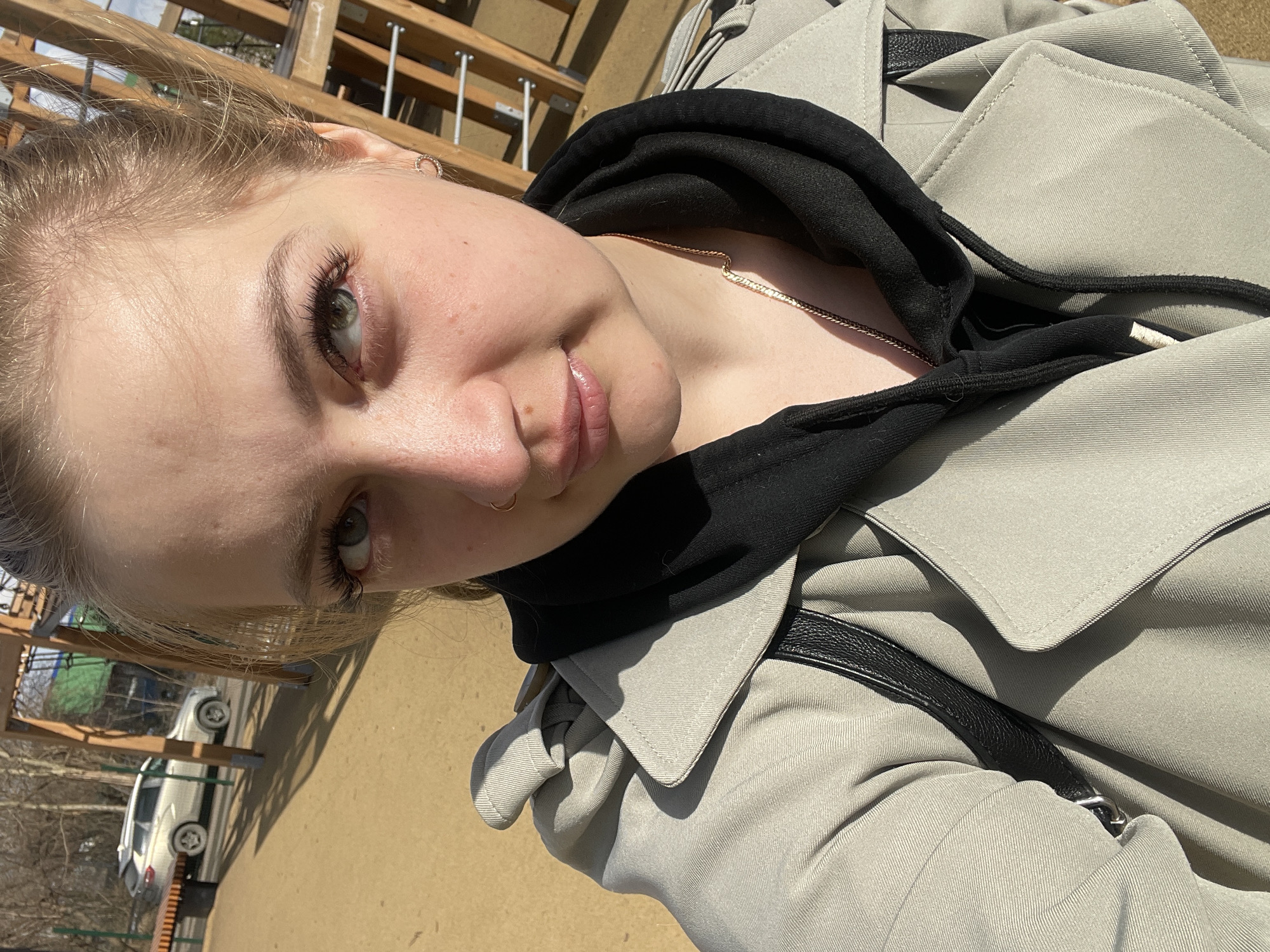 The image shows a selfie of a young woman with light blonde hair and fair skin. She has green eyes and is wearing a black hoodie under a light gray trench coat. A thin gold necklace is visible around her neck, and she has a nose ring. The photo is taken outdoors in sunlight. In the background, there's a wooden structure, possibly part of a playground or park facility. Lower in the background, cars are visible, and there is possibly some grass or greenery. There's nothing specifically that confirms the location as the city of Izhevsk, Russia.