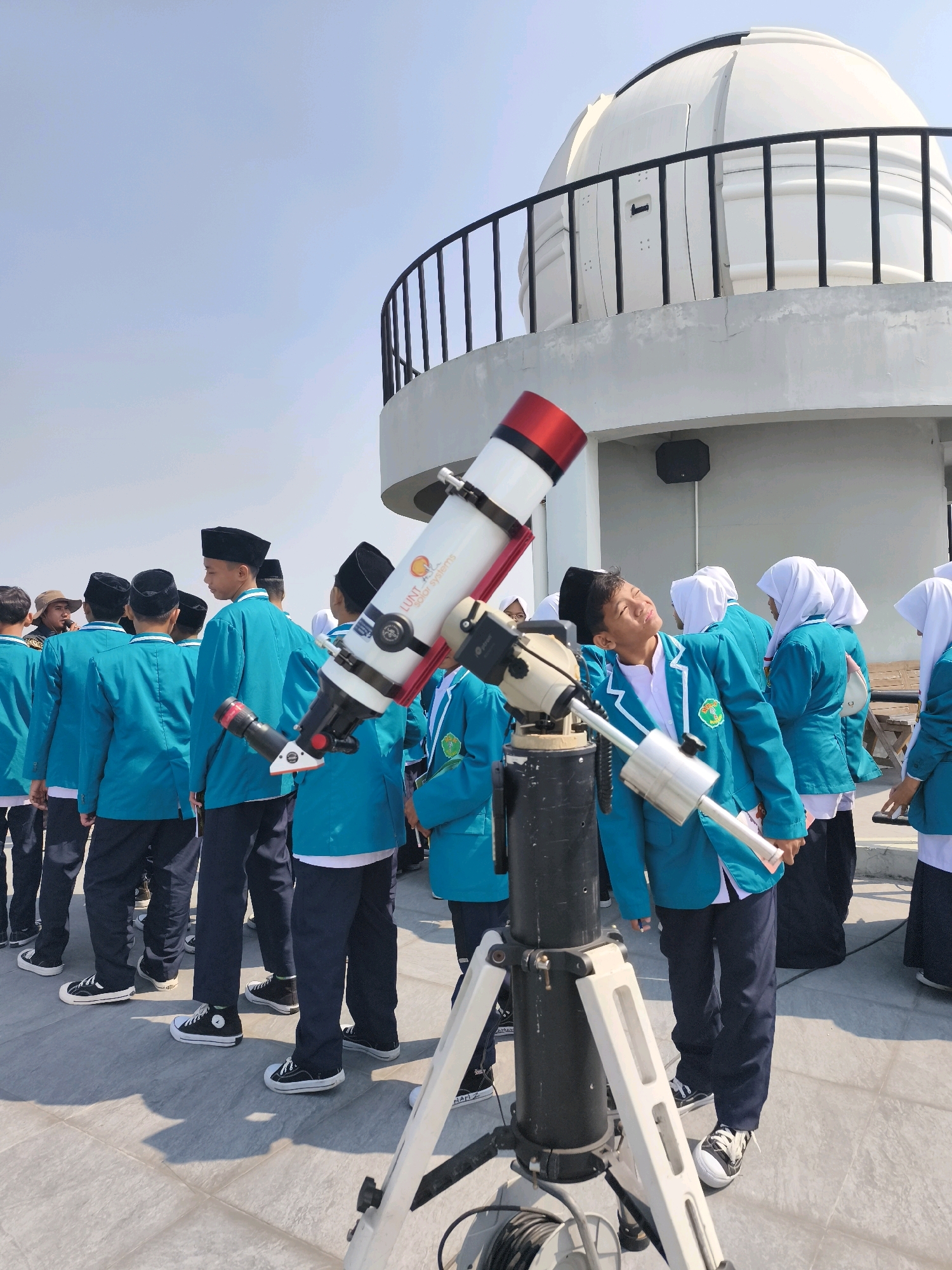 A group of students, likely on a field trip, are gathered on the rooftop of an observatory in Semarang, Indonesia. The observatory, a dome-shaped structure, is visible in the background. In the foreground, a telescope is set up on a tripod, with one student looking through it. The students are wearing matching turquoise jackets and dark pants, with many also wearing black skullcaps. The sky is clear and blue, suggesting a sunny day. The students appear engaged and curious, with some pointing and discussing the telescope. The overall scene conveys a sense of educational exploration and scientific interest.