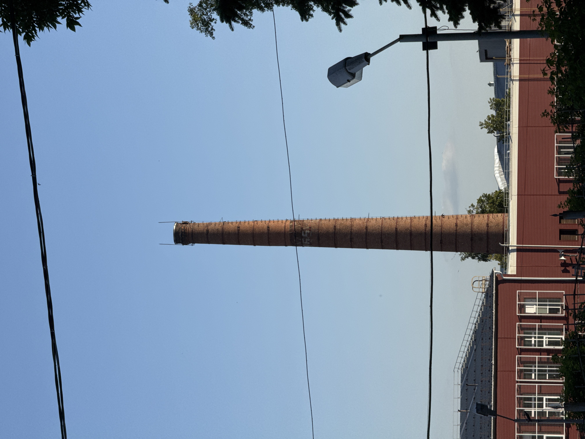 An outdoor, eye-level, medium shot shows a tall, brick smokestack on a clear, blue sky day. The smokestack, located in Izhevsk, Russia, is brick-red and appears to be old. It has a metal ladder running up its side and a ring of metal at its top. The smokestack is in the center of the frame and is the dominant subject. In the foreground, a red, corrugated metal building with several windows and a light post are visible. Power lines stretch across the sky, obscuring parts of the smokestack and building. Trees are visible on the right side of the image. The scene suggests an industrial or manufacturing setting.
