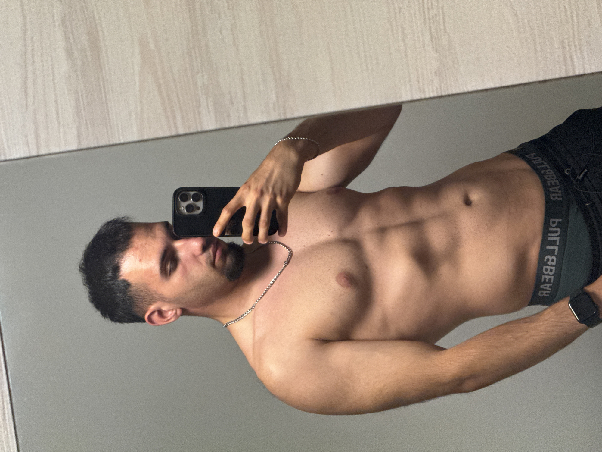 A shirtless man with dark hair and a goatee is taking a mirror selfie. He is wearing black athletic shorts with a logo waistband and a silver chain necklace. He has a watch on his left wrist. The background appears to be a bathroom with a tiled wall and a grey wall. There is no indication of the city of Shumen, Bulgaria in the image.