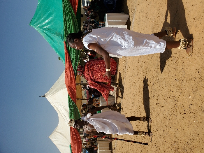 The scene depicts an outdoor gathering in Bar Gada, Nigeria, under a clear blue sky with bright sunlight. The foreground features two barefoot individuals on reddish-brown, sandy ground. On the left, an adult male with dark skin wears a white loincloth, an ornate light-colored headdress, and light-colored anklets. He holds a multi-banded staff vertically in his left hand and faces right. On the right, an adult female with dark skin wears a short-sleeved white tunic dress, light-colored anklets, and wrist bracelets. She extends both arms forward, manipulating or presenting a large, textured red fabric. In the background, multiple white and green peaked tents provide canopy cover, with additional red and green fabric drapes strung horizontally. Numerous attendees are visible under the tents, some seated at white-clothed tables. Shadows cast by the foreground figures are distinct on the ground.
