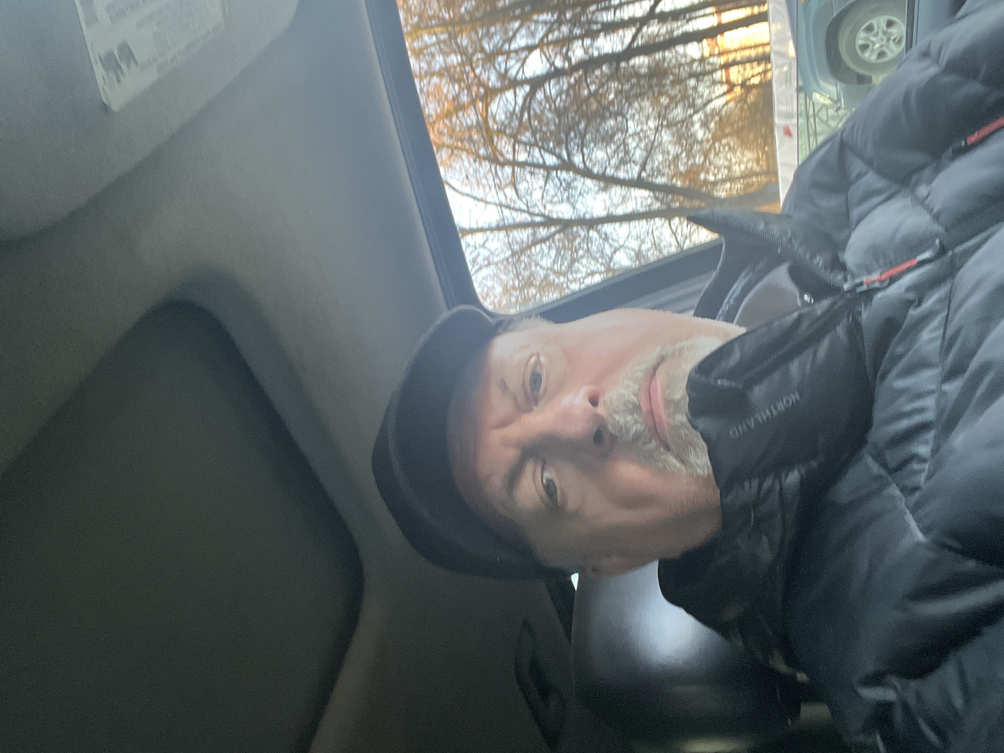 A man is visible in the photo, wearing a dark-colored cap and a puffer jacket. The jacket has a label visible near the neck area. The man is inside a vehicle with a sunroof, through which trees and another vehicle are visible. The interior of the car is dark-toned. The image does not allow for the identification of Podolsk, Russia.