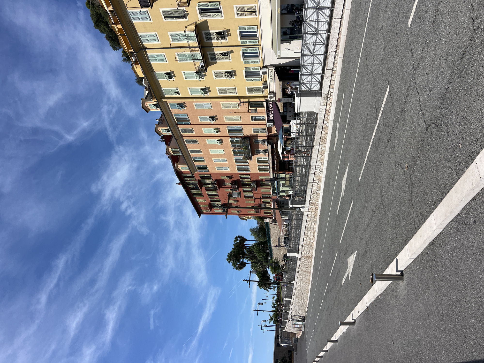 This is a photograph taken from street level, looking down a street lined with buildings in Nice, France, specifically along the Promenade des Anglais. The sky is a vibrant blue with wispy white clouds, and there are visible contrails from airplanes.
In the foreground, a two-lane asphalt road curves gently to the left. White lane markings and a large white arrow indicating traffic direction are visible on the road. The left side of the road is bordered by a concrete curb and a line of bollards.
Adjacent to the road is a raised sidewalk or promenade area. This area is separated from the road by a low stone wall and a decorative metal railing. Behind the railing, several people are walking and sitting at outdoor cafe tables. Palm trees are visible in the background, contributing to the Mediterranean atmosphere.
The main subject of the photograph is a row of colorful buildings lining the promenade. The buildings are tall and narrow, painted in shades of red, orange, yellow, and cream. They feature multiple stories with numerous windows, many of which have light-colored shutters, some open and some closed. Small balconies with black railings are also present on some of the buildings. The architectural style suggests a European, possibly French, influence.
On the ground floor of the buildings, several shops and restaurants are visible with awnings and signage. One sign reads "NICE DOCK". The buildings appear to be situated on a slight incline, with greenery and trees visible on the hillside behind them. The overall scene depicts a bright, sunny day in a coastal city.